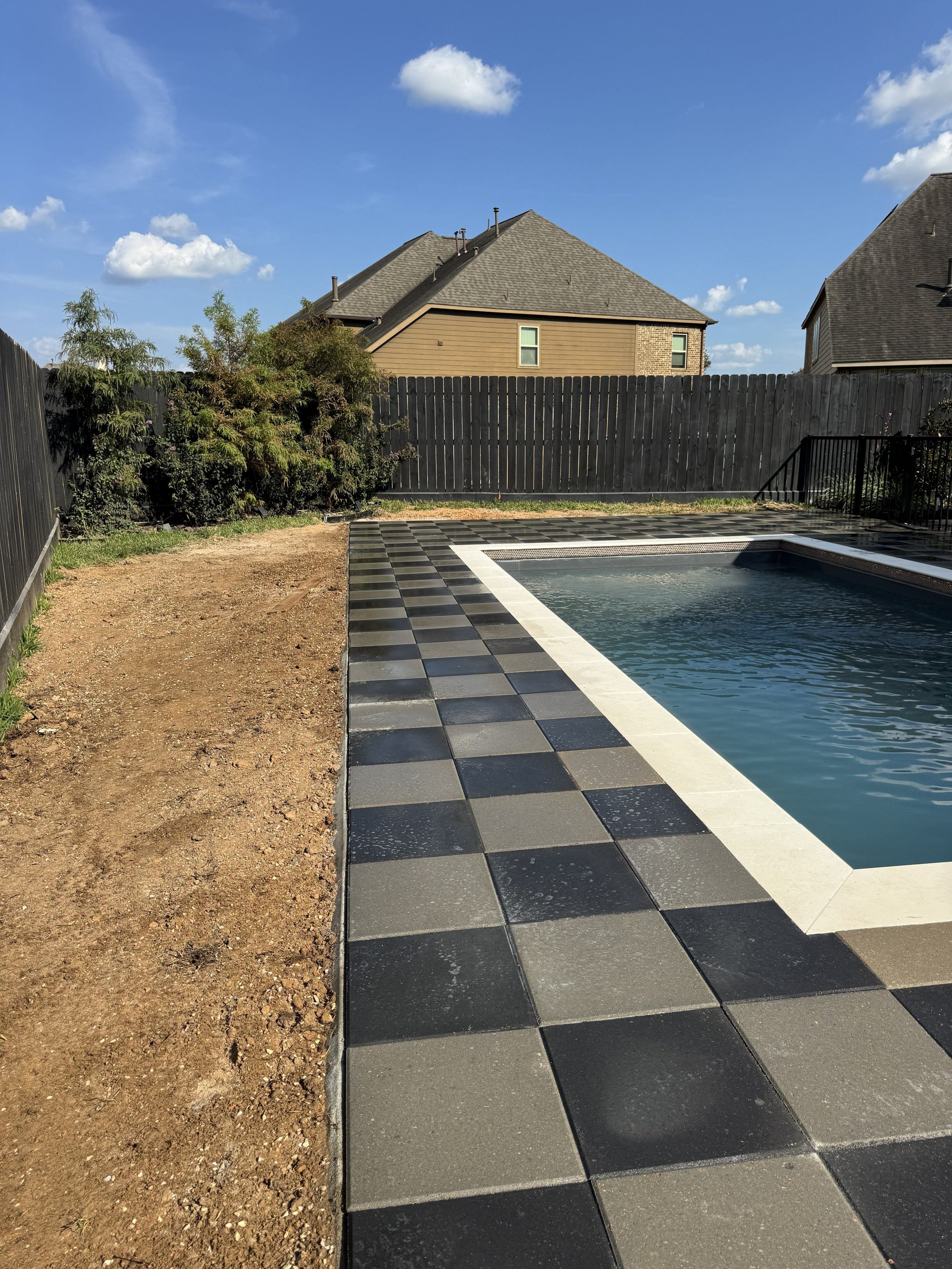 Backyard with a small swimming pool, checkered black and gray pool deck, and a black wooden fence, with houses and blue sky in the background.