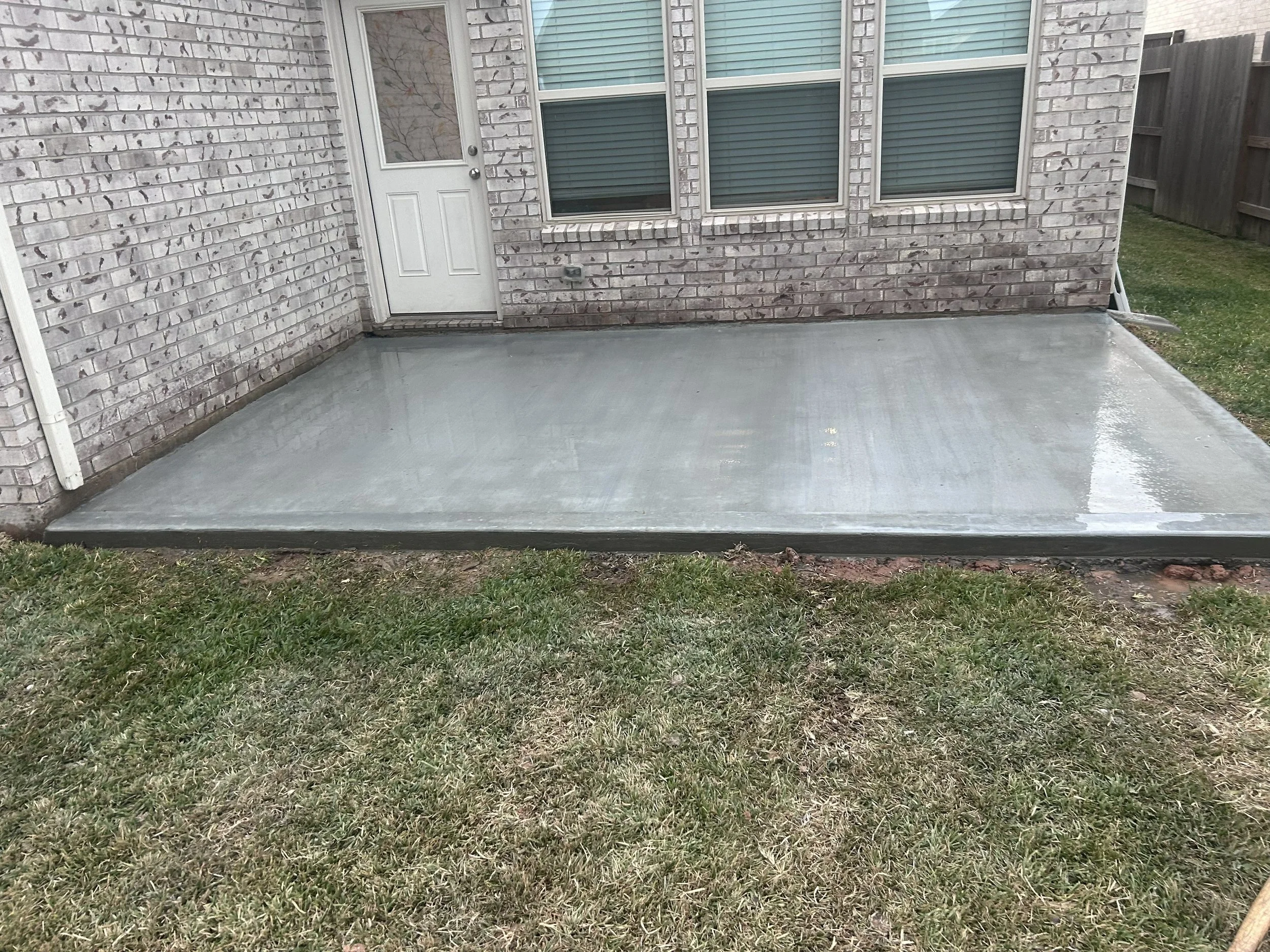 Backyard patio with freshly poured gray concrete, surrounded by a grass lawn and brick house exterior with three windows and a door.