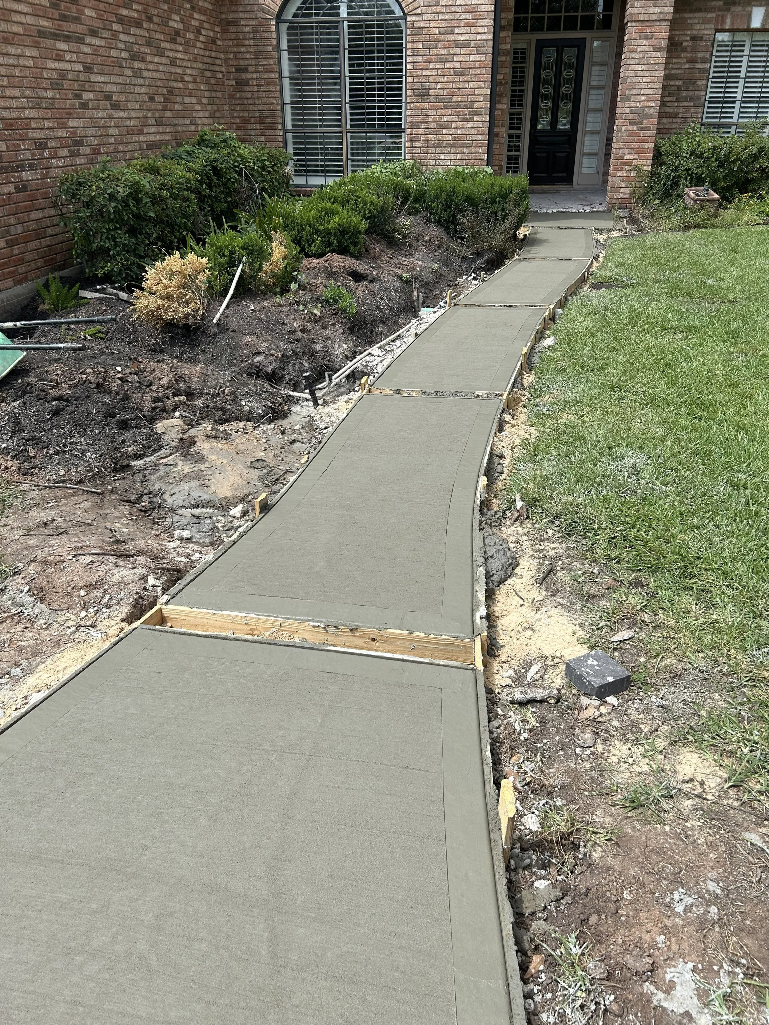 Concrete sidewalk being constructed in front yard of brick house, with part of the old dirt and grass area still visible.