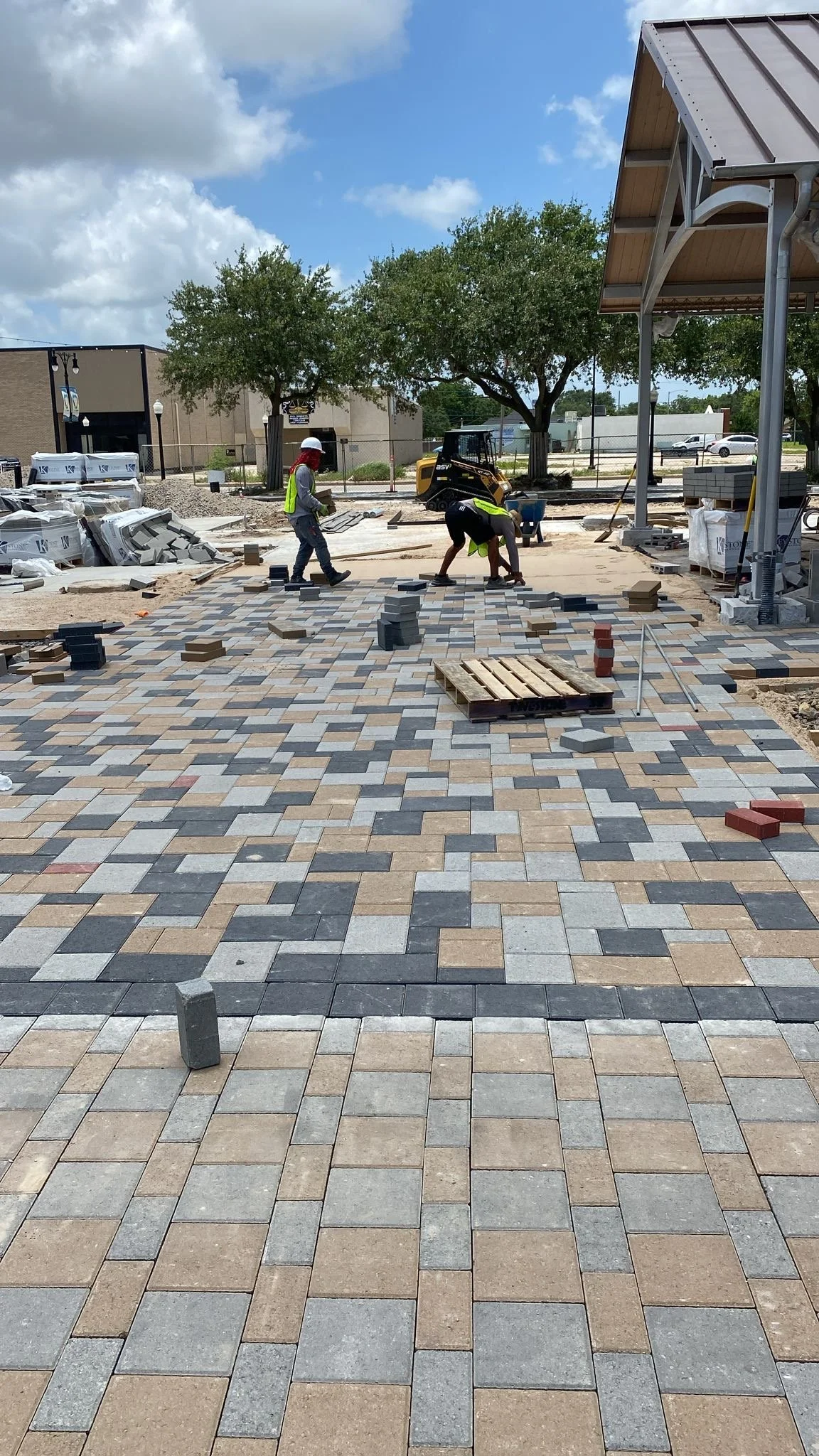 Workers laying down paving stones to create a patterned walkway outdoors, under a partly cloudy sky, with trees and construction equipment visible in the background.