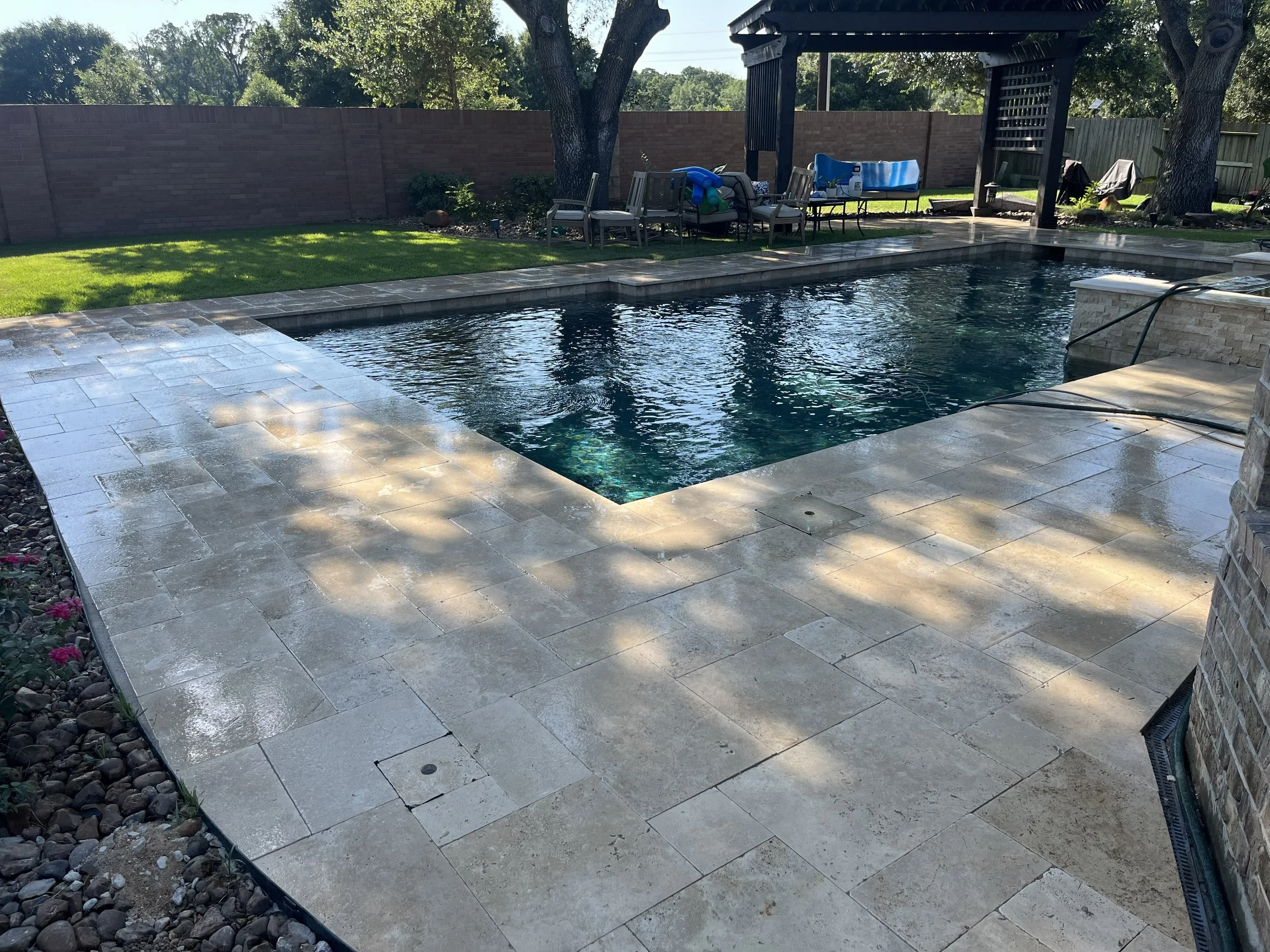 A backyard swimming pool area with paved stone deck, surrounded by green grass, trees, and a brick fence. There are chairs, a table, and a covered seating area in the background.