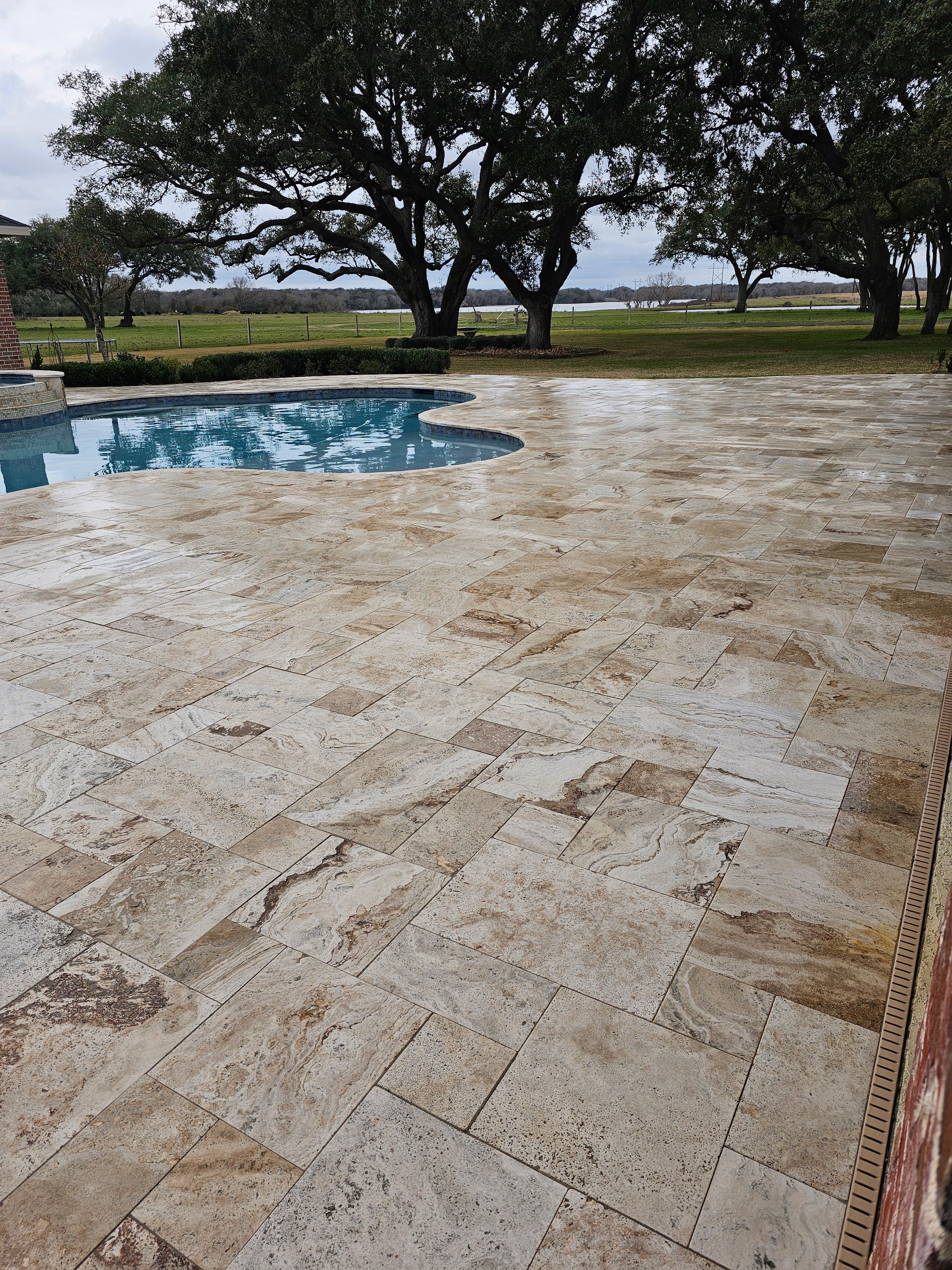 A pool on a stone patio with trees and a field in the background.