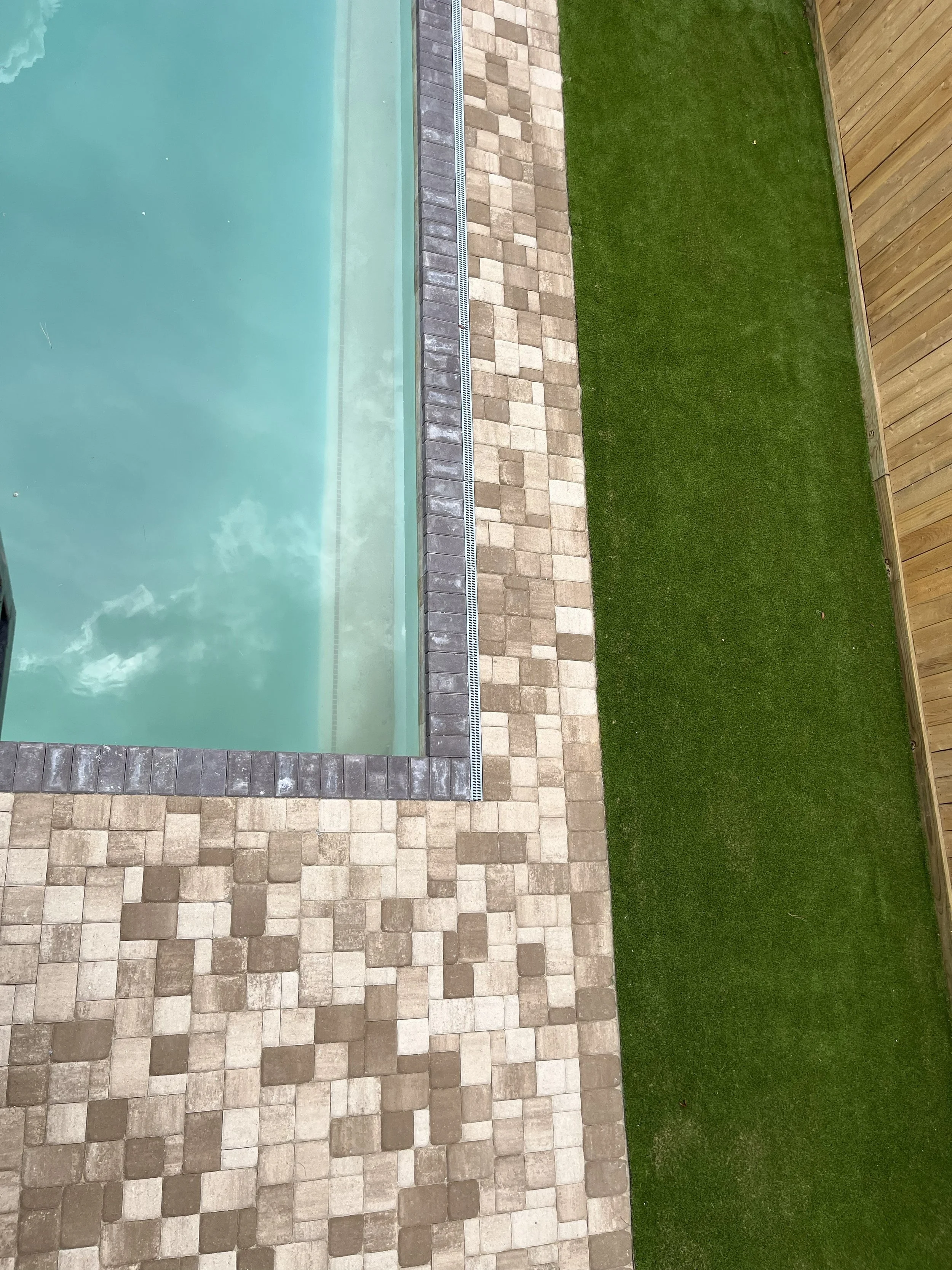 Backyard with a swimming pool, a brick and stone patio, and a wooden fence with a green grassy lawn.