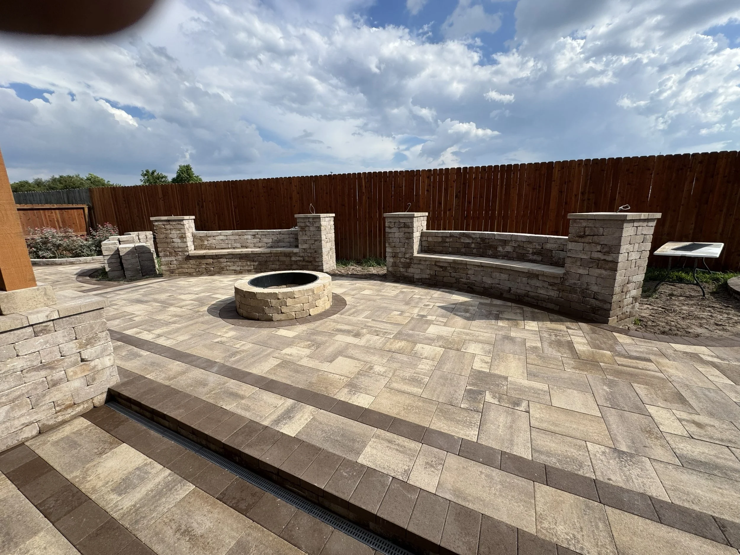 An outdoor patio with a curved brick fire pit in the center, surrounded by a low brick wall, with decorative bushes and a wooden fence against a cloudy sky.