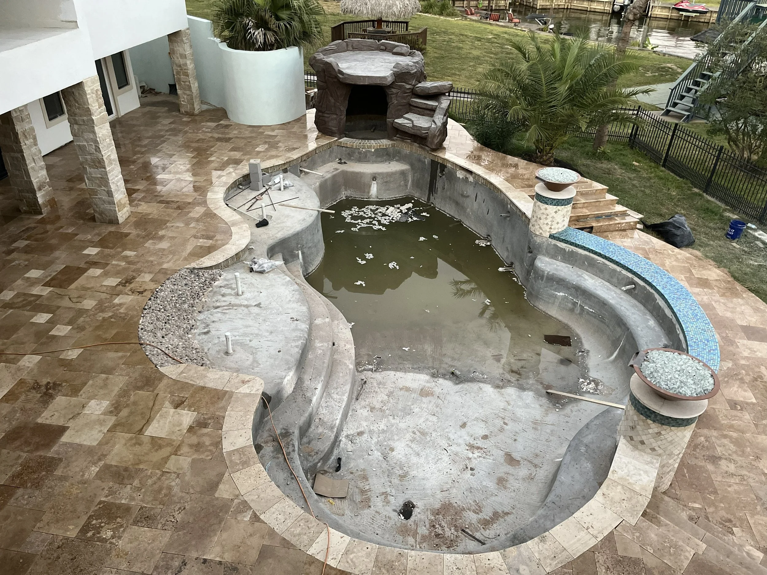 Empty swimming pool under construction with dirty water and construction tools around, patio area with brown tile flooring, outdoor fireplace, and a view of a backyard with grass, trees, and a canal in the background.