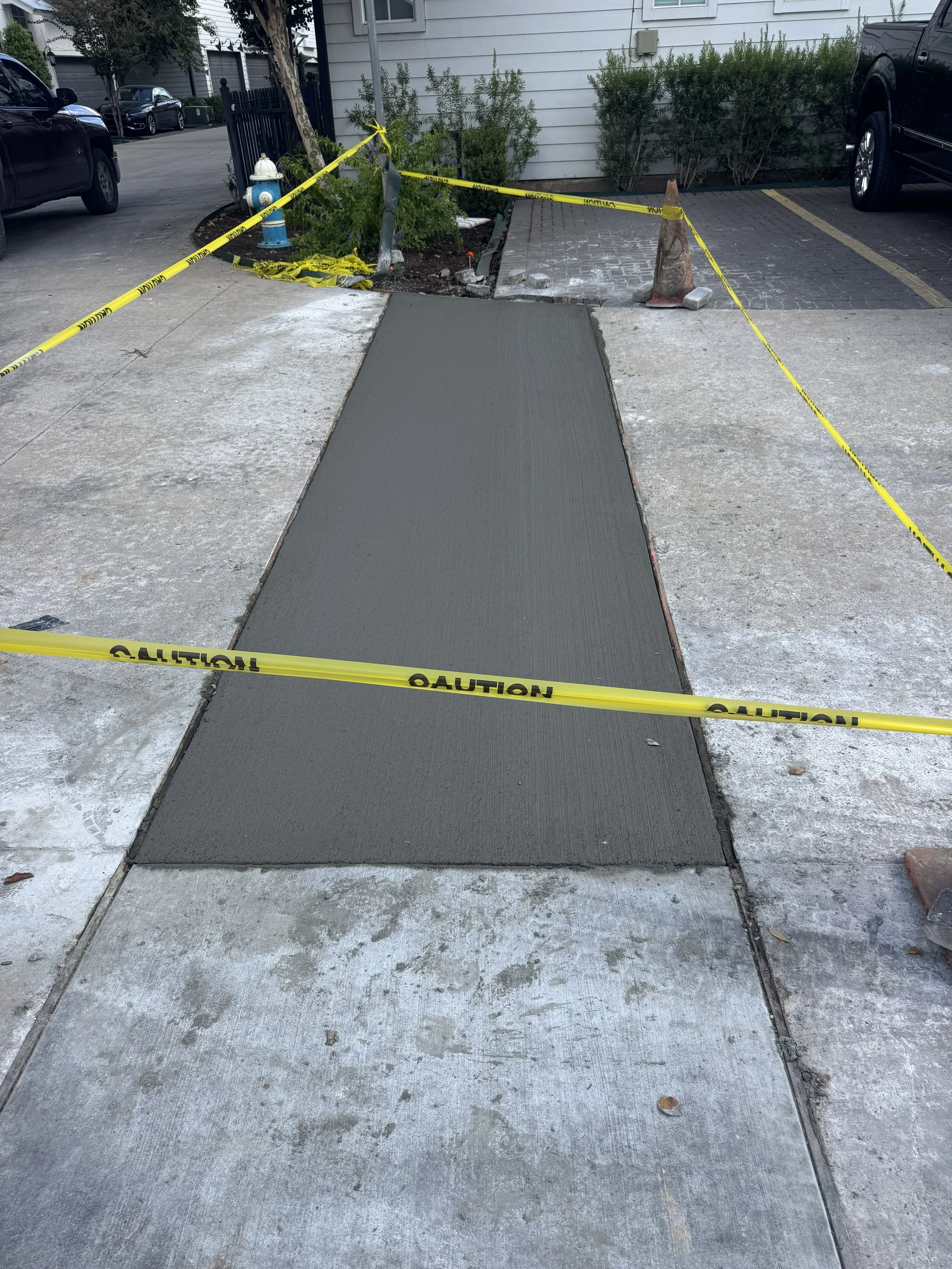 Freshly poured concrete on a sidewalk section, with caution tape and traffic cones surrounding the area, while parked cars and residential buildings are visible in the background.