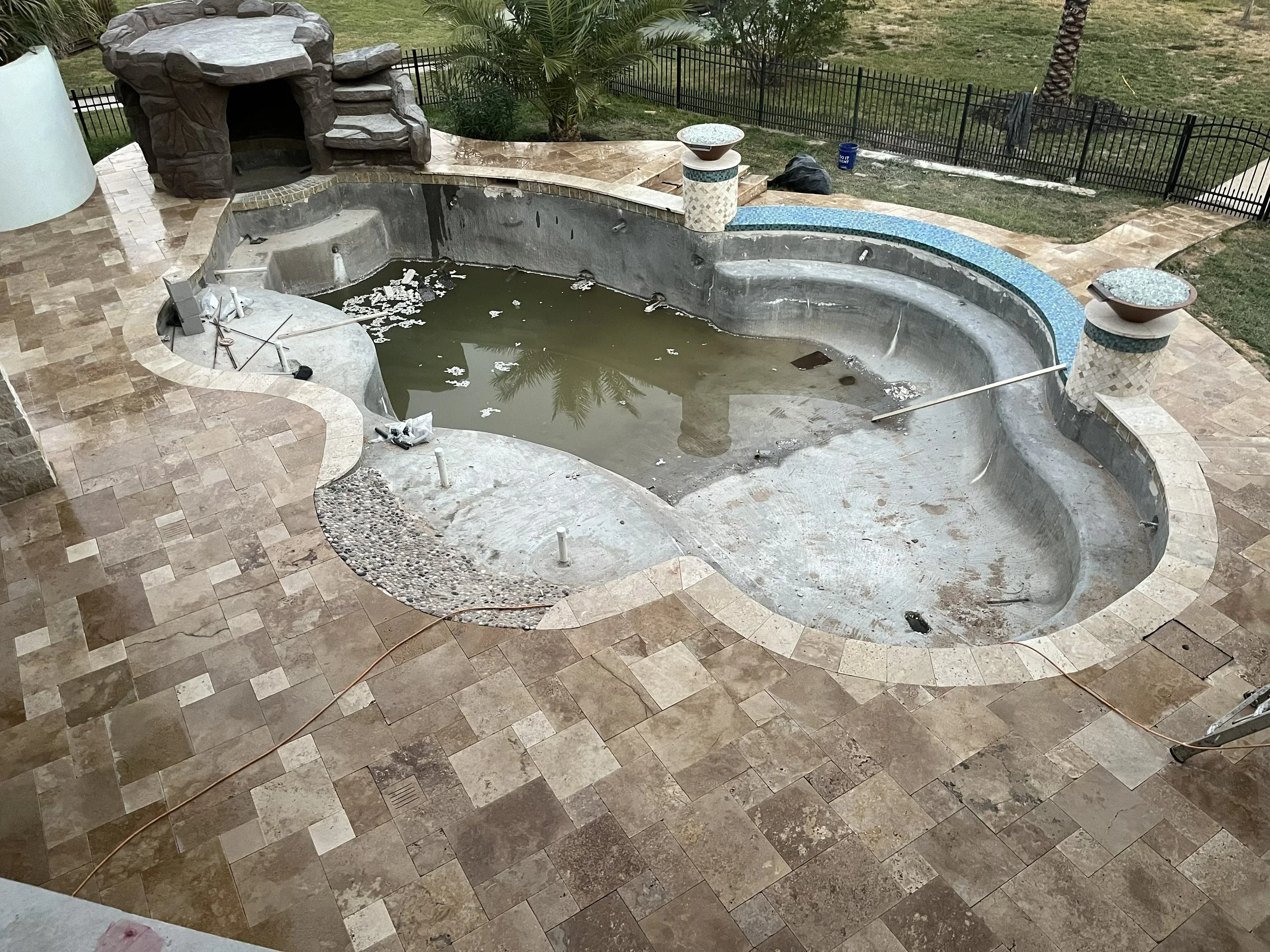 Empty in-ground swimming pool with dirty water, debris, and construction materials, surrounded by a stone tiled deck and a black metal fence in a backyard.