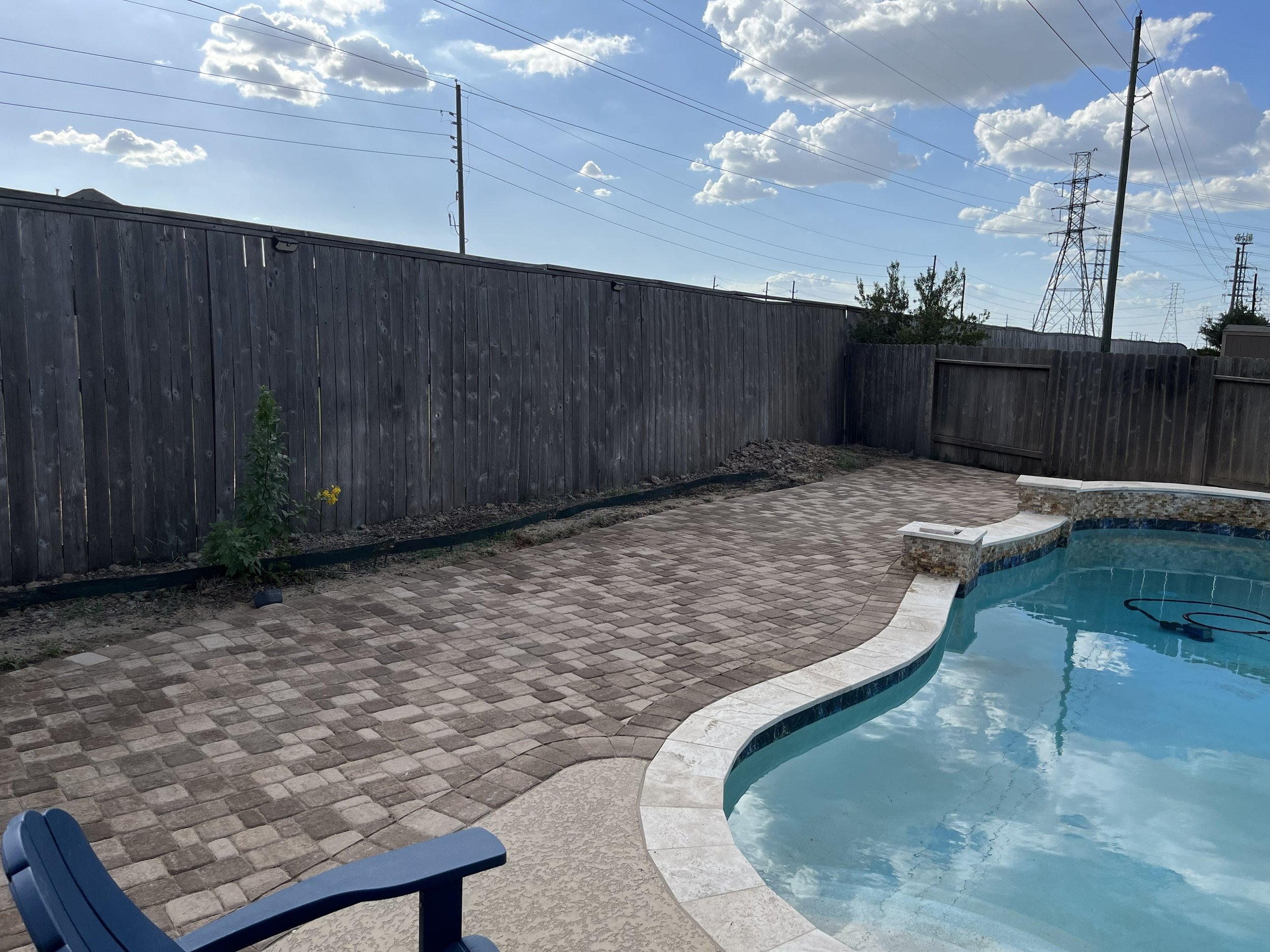 A backyard swimming pool with a curved edge, surrounded by a brick patio. A wooden privacy fence and power lines with utility poles are visible in the background. The sky is partly cloudy with blue sky and white clouds.