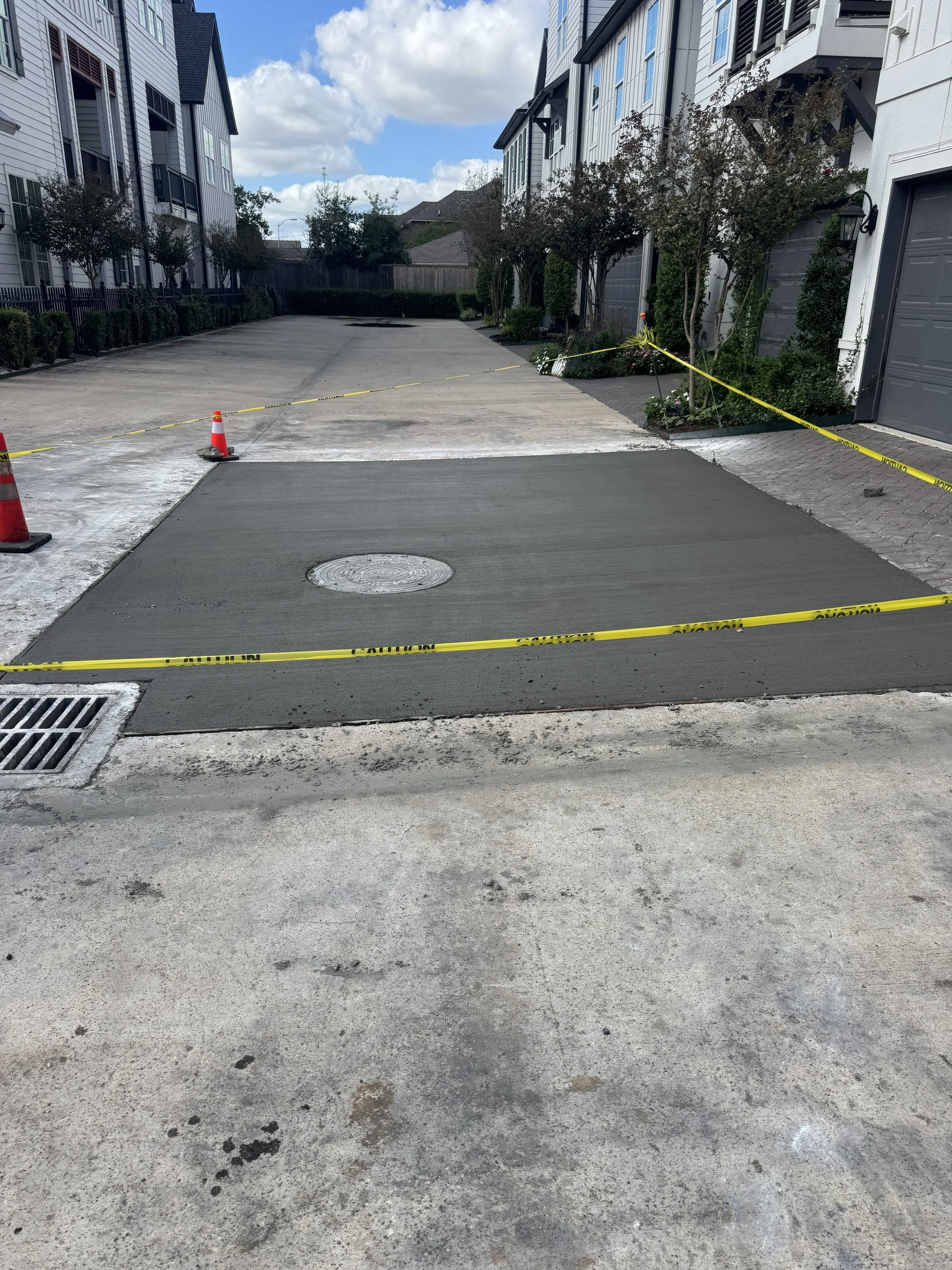 A residential driveway with fresh poured concrete section, surrounded by caution tape and traffic cones, with apartment buildings on either side and a cloudy sky overhead.