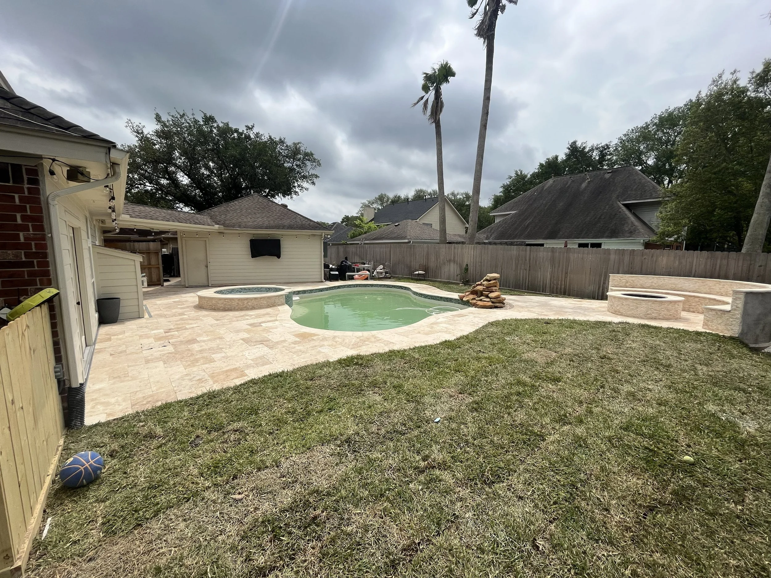 A backyard with a swimming pool, a hot tub, and a grassy area with a basketball on the ground. There are trees and houses in the background under a cloudy sky.
