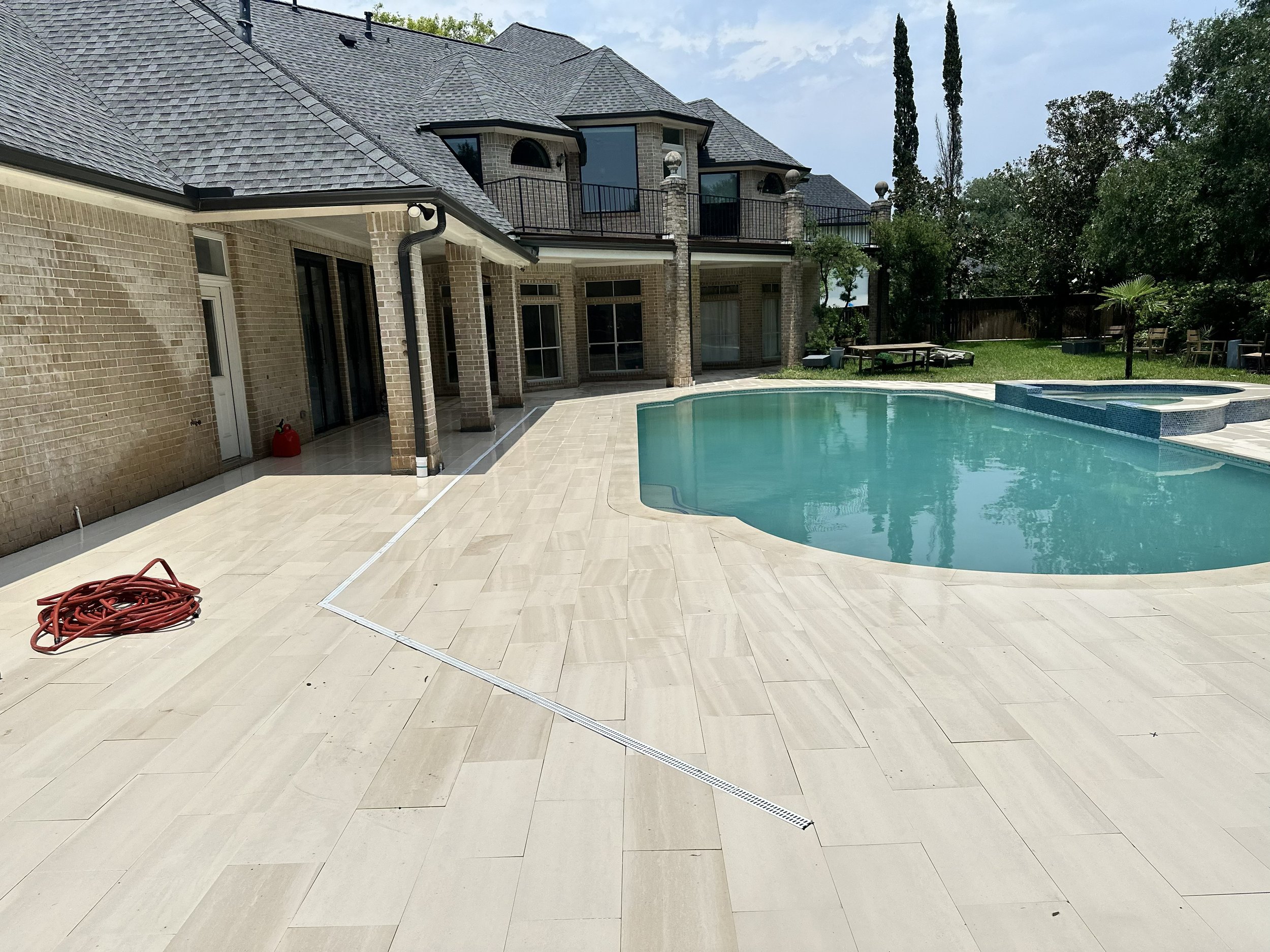 A backyard with a swimming pool, a tiled patio, and a multi-story brick house with a balcony and grey roof. There are trees, outdoor furniture, and lawn area, with a coiled red garden hose on the patio.