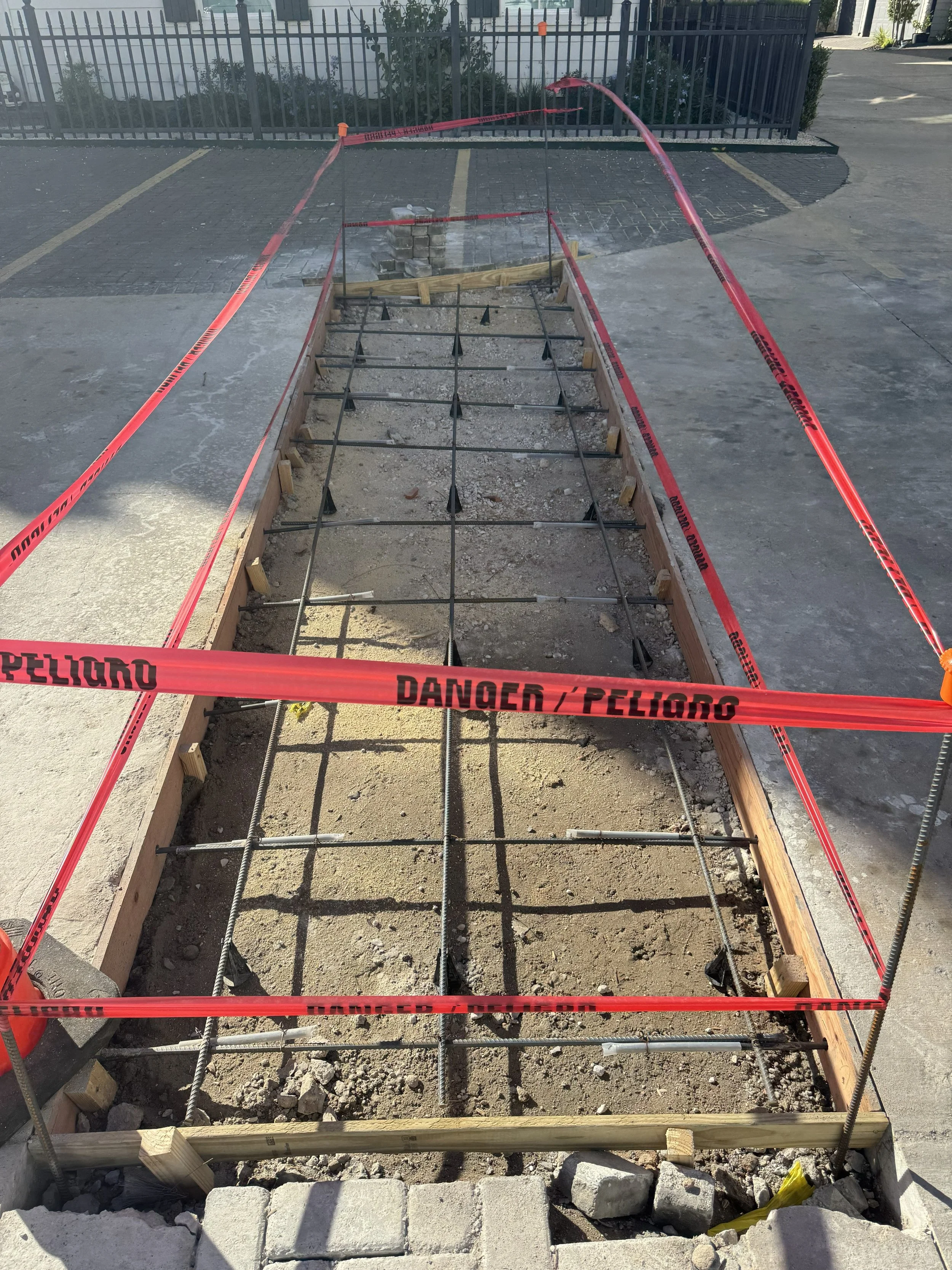 A construction site for a sidewalk or pathway with a wooden frame and steel reinforcement bars, surrounded by red 'DANGER / PELIGRO' tape, with a paved parking lot and fence in the background.