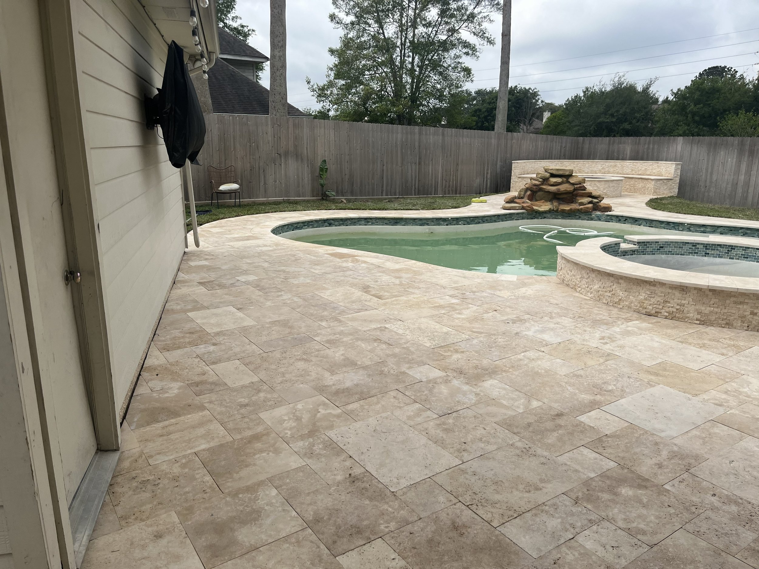 Patio area with beige tiles, a corner of a house wall on the left, a door with a black bag hanging on it, a wooden fence enclosing an outdoor pool with a rock waterfall feature, a chair with a white cushion, and trees in the background under cloudy s