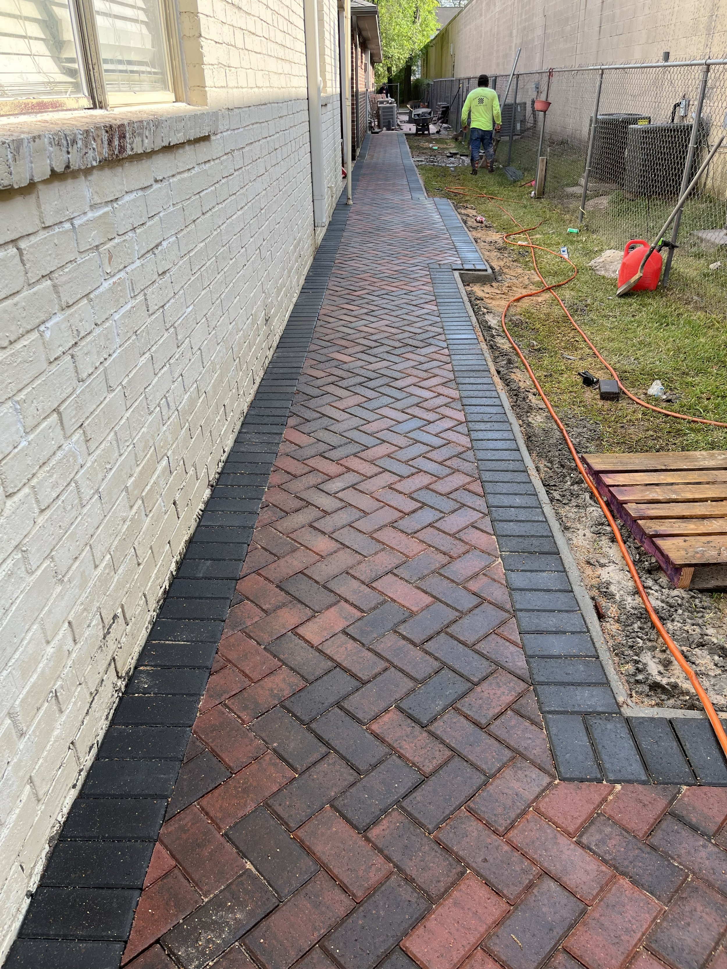 A newly paved brick sidewalk running alongside a white brick building with a window on the left. The sidewalk is bordered by black bricks along the edges. In the background, two workers are working near a fence, with construction tools and equipment 