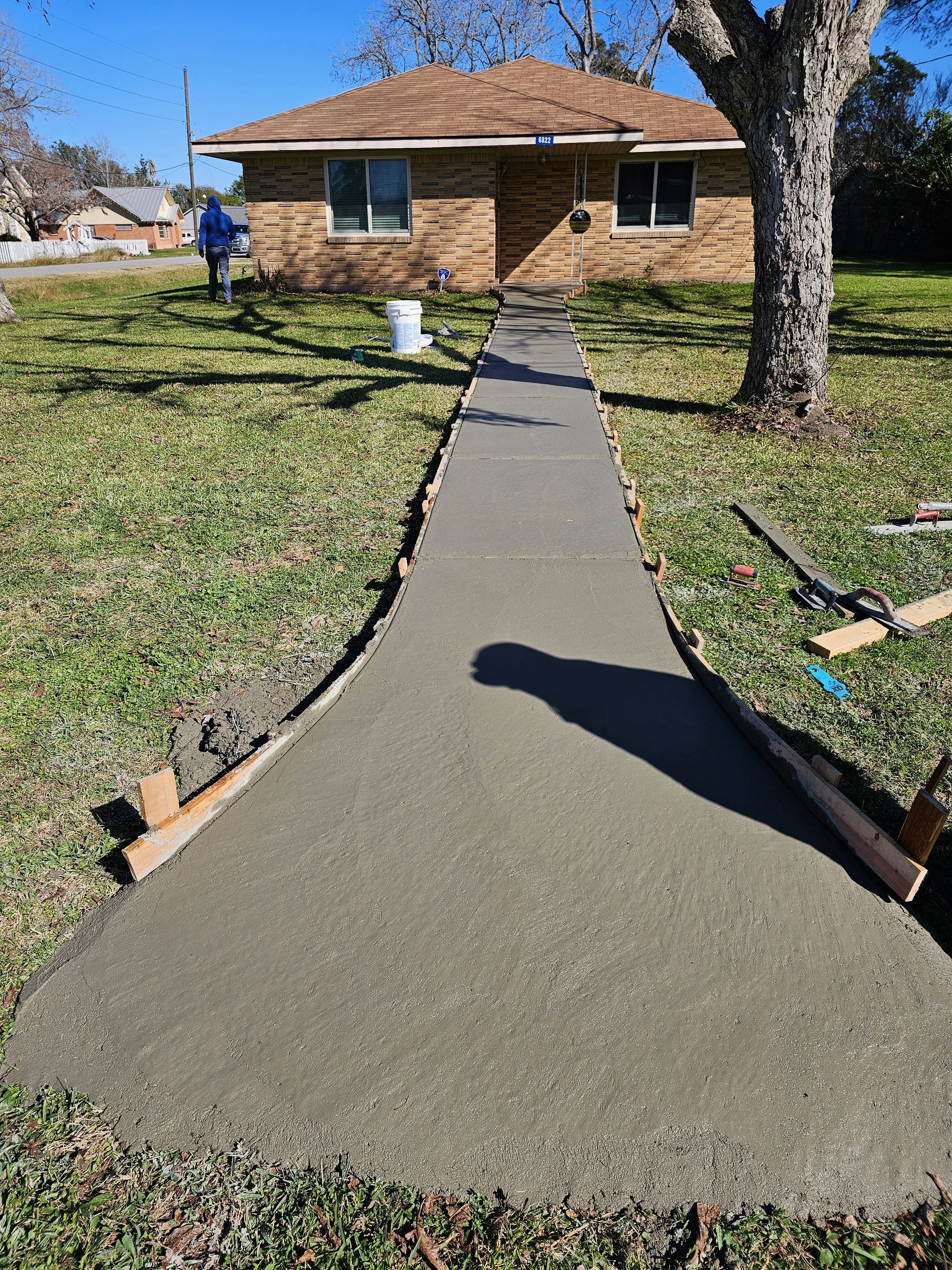 Newly poured concrete sidewalk leading to a brick house with a brown roof, shaded by trees, with a person in a blue hoodie walking away in the yard.