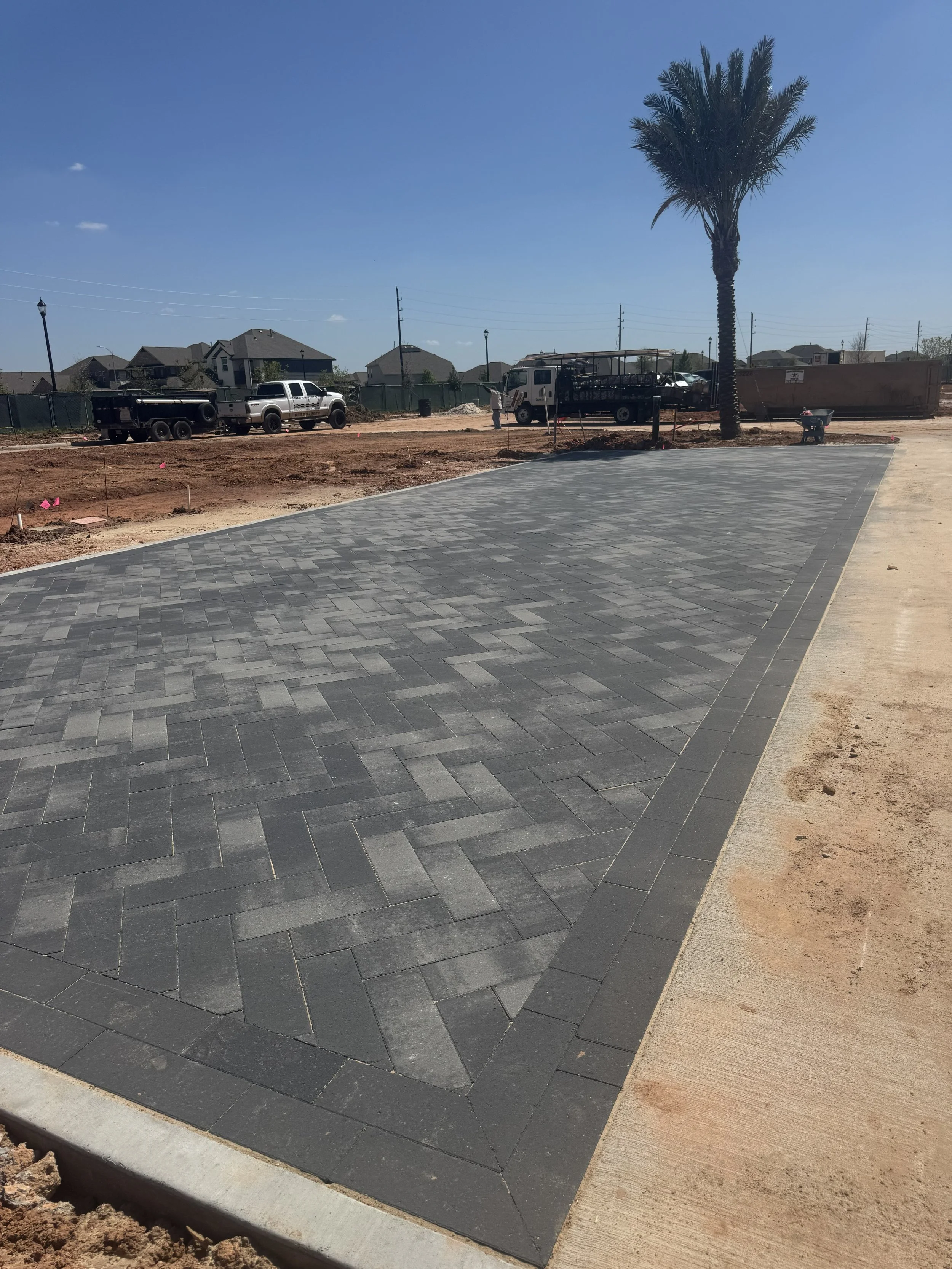 A newly paved sidewalk with gray brick pavers next to a construction site with trucks, a palm tree, and houses in the background under a clear blue sky.