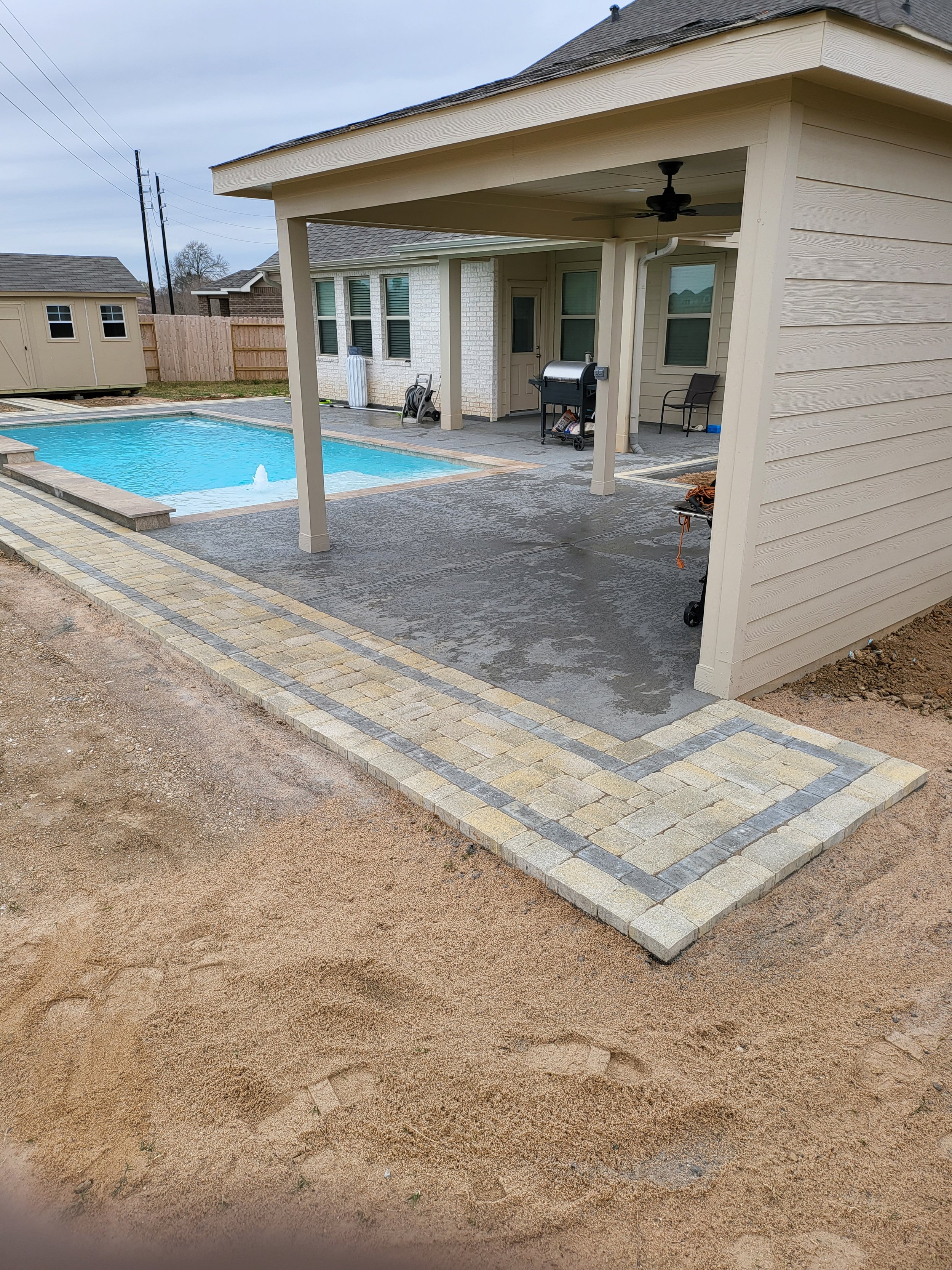 A backyard with a swimming pool, a covered patio area with a ceiling fan, a grill, and a chair. The ground is partially paved with bricks and partially dirt.