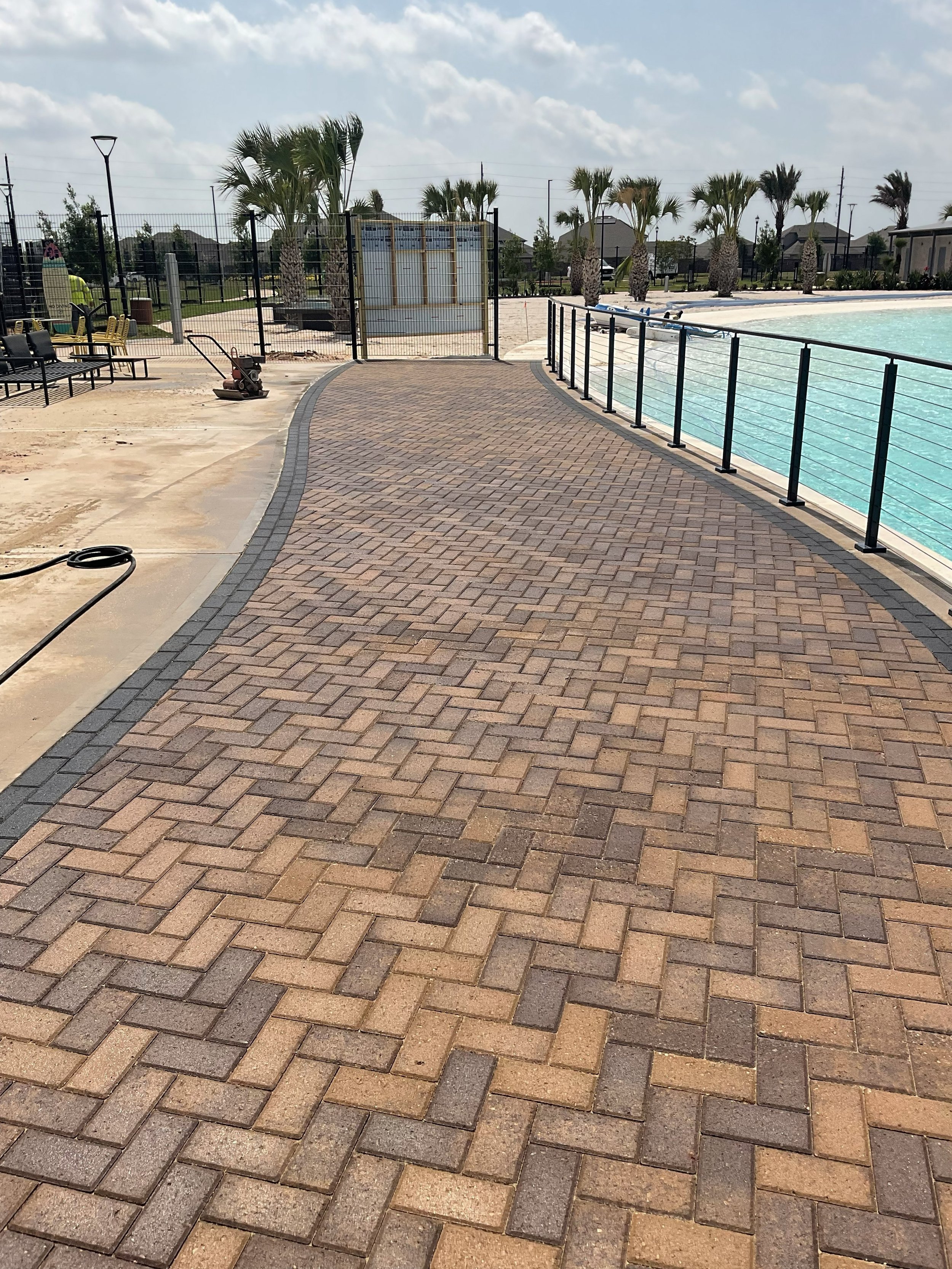 A paved walkway next to a swimming pool with water, black metal fencing, palm trees and houses in the background, and a few workers and equipment.