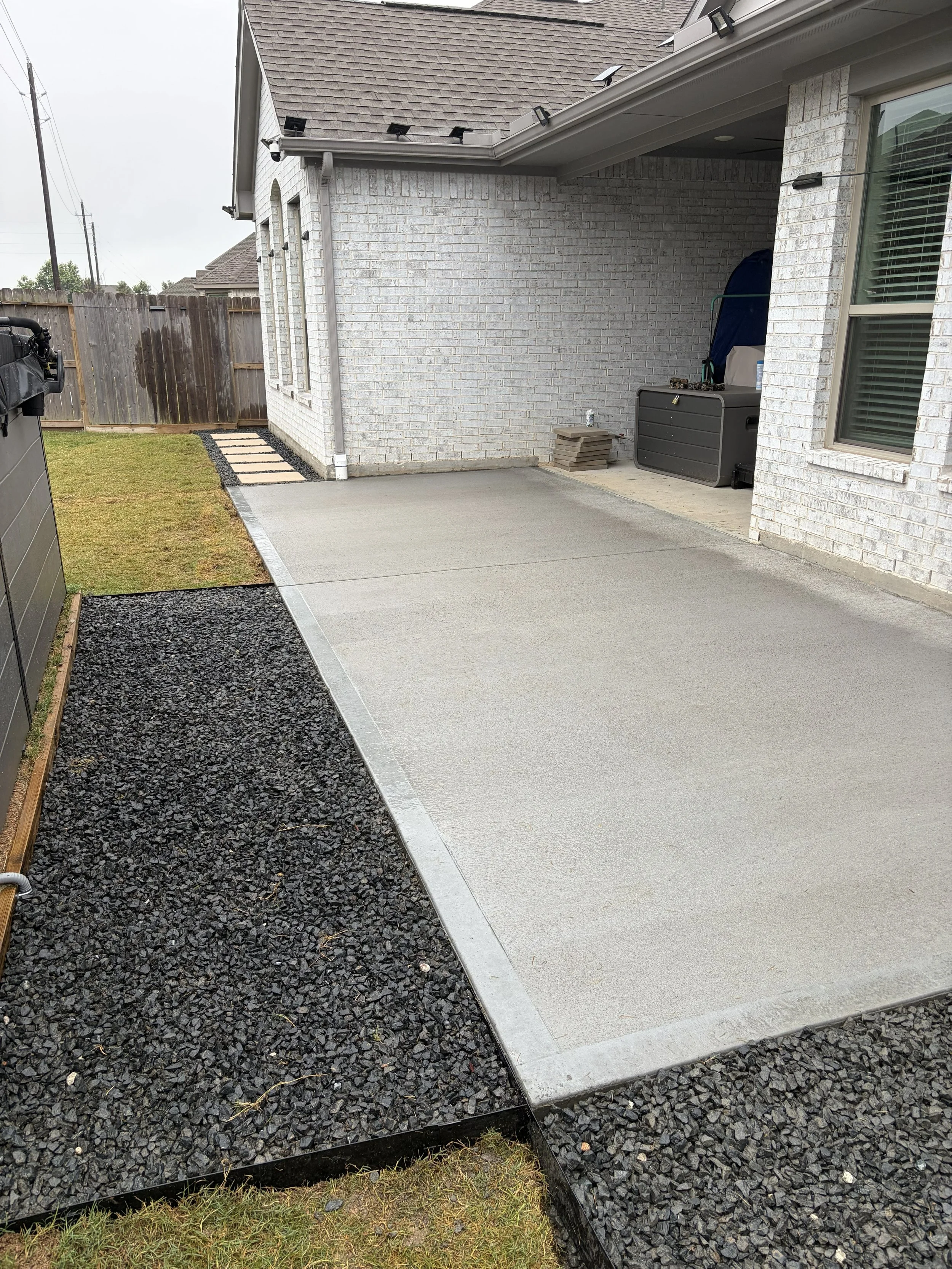 Backyard patio with a concrete area, black gravel border, a grassy section, and a wooden fence. The house has white brick walls, multiple outdoor lights, and a window with blinds. There is a small patio storage bin, some stacked pavers, and a propane