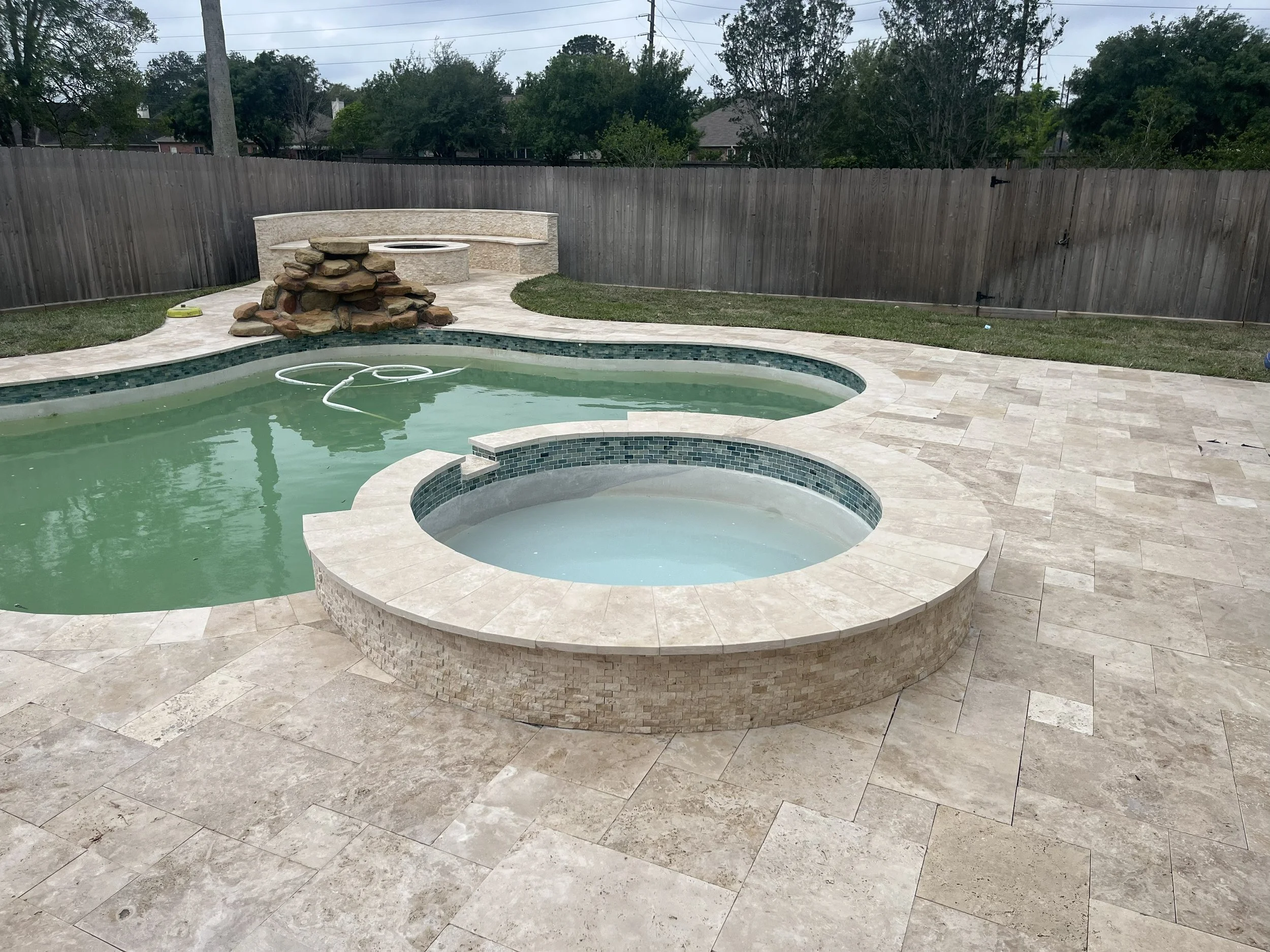 Backyard with a swimming pool, hot tub, and a fire pit made of stacked rocks, surrounded by a wooden fence and trees.