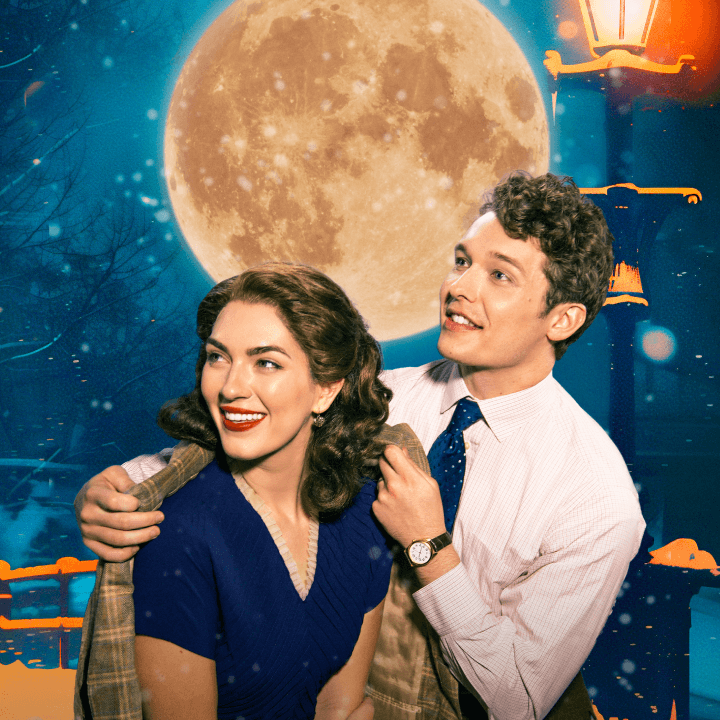 George and Mary from It’s a Wonderful Life stand together outdoors at night as snow falls, illuminated by a glowing streetlamp and a large full moon.