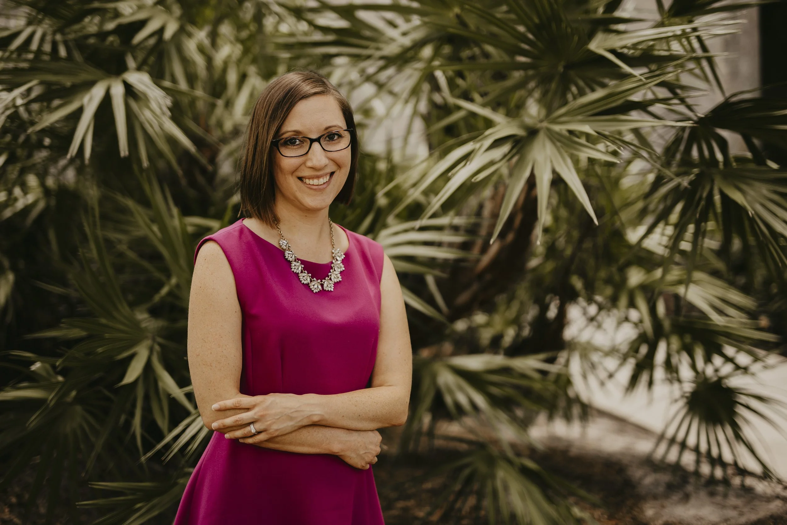 A woman wearing a fuchsia sleeveless dress and a statement necklace stands outdoors in front of a large leafy plant, smiling at the camera.