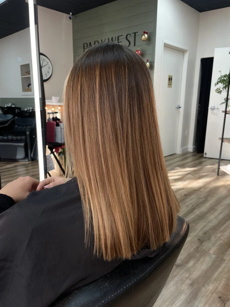 Back view of a woman with straight, shoulder-length hair a mix of light brown and caramel highlights, sitting in a salon chair.
