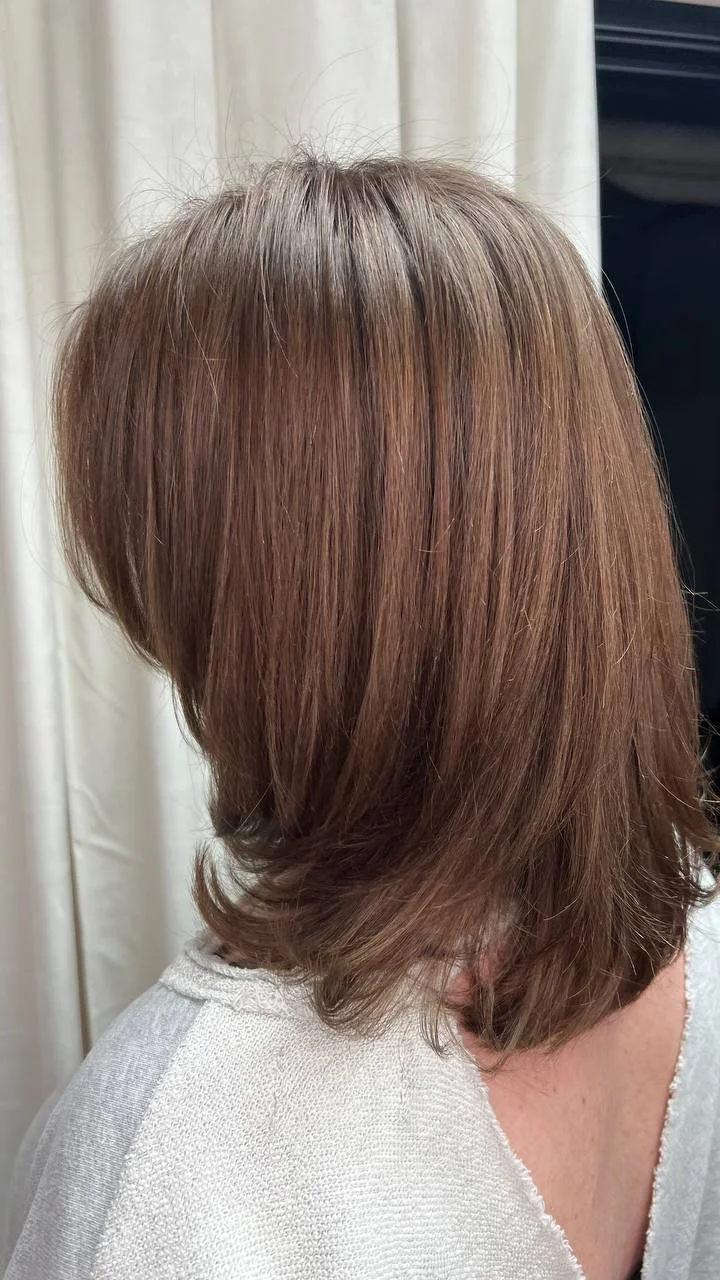Back of a woman's head with medium-length brown hair, layered and slightly curled at the ends, standing in front of a light-colored curtain.