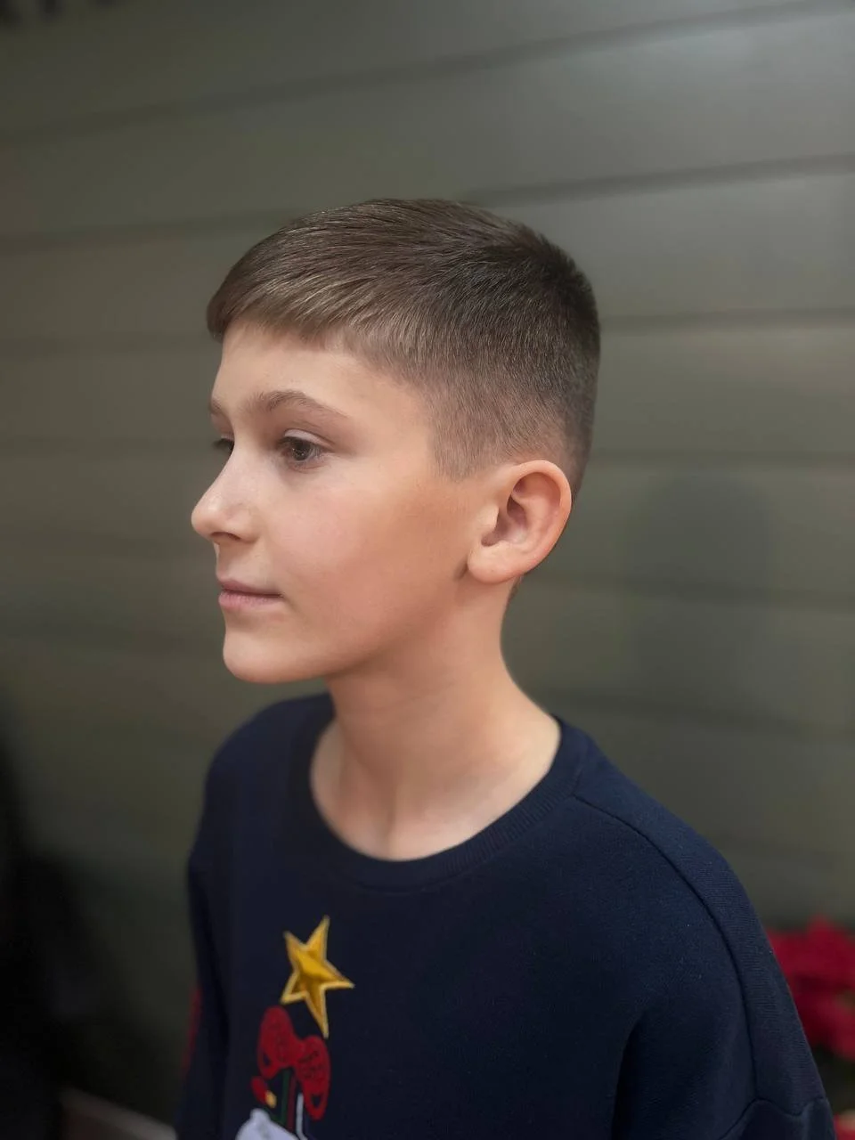 Side profile of a young boy with a short, fade hairstyle wearing a navy blue shirt with a star and cupcake graphic, standing in front of a gray wall.