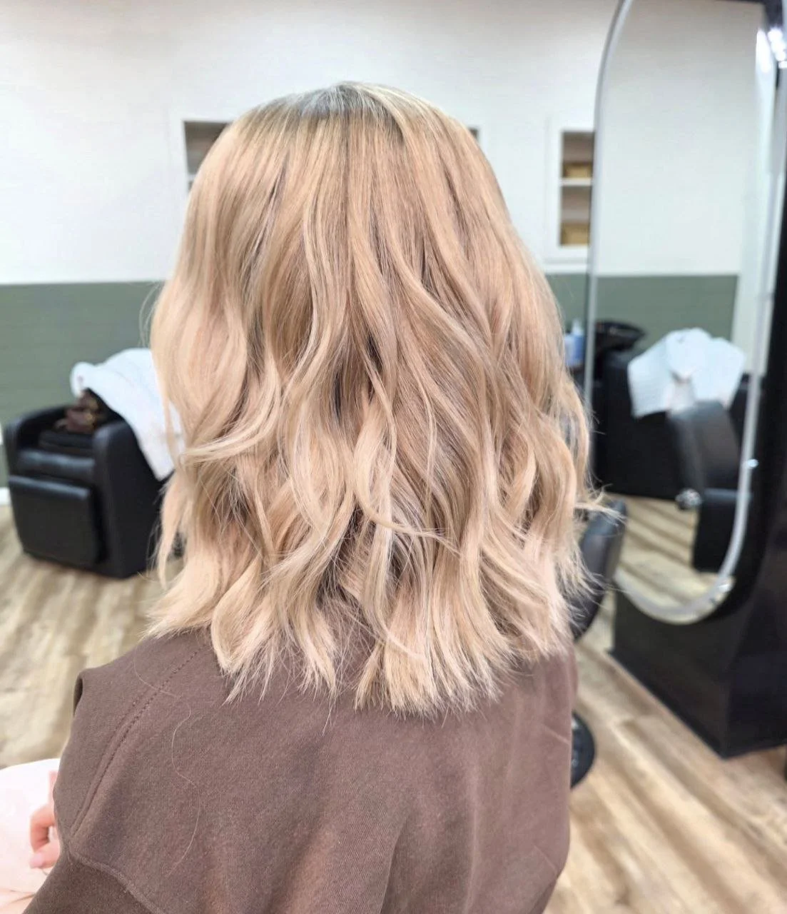 Back view of a woman with shoulder-length wavy blonde hair in a salon with a mirror and chairs.