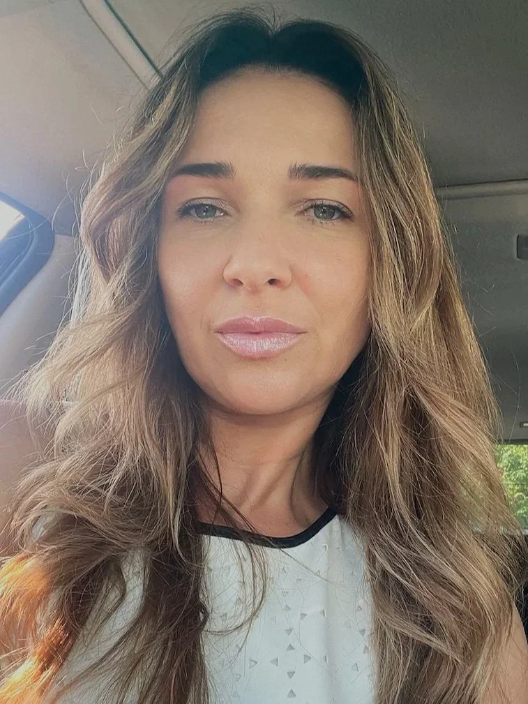 Close-up of a woman with long, wavy brown hair and light makeup, sitting inside a vehicle with sunlight illuminating her face.