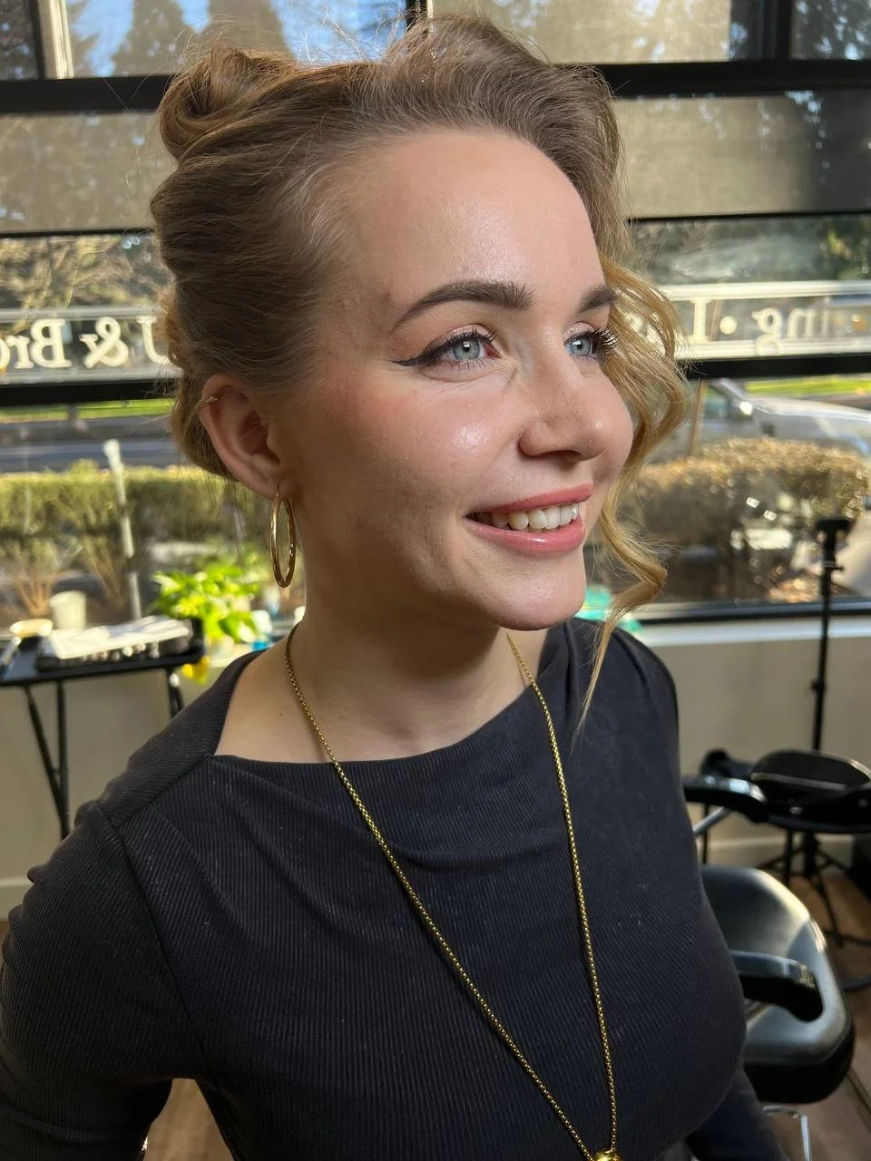 A smiling woman with blonde hair styled in loose waves, wearing a black top, gold hoop earrings, and a gold necklace, standing indoors with sunlight coming through a window behind her.