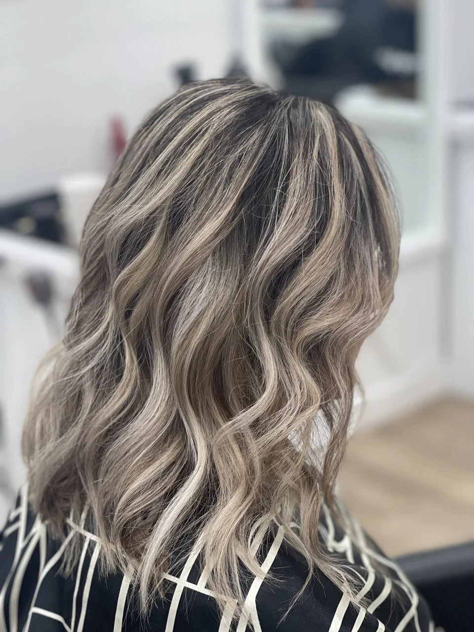 Side view of a woman with shoulder-length, wavy blonde hair with darker roots, sitting in a salon chair.
