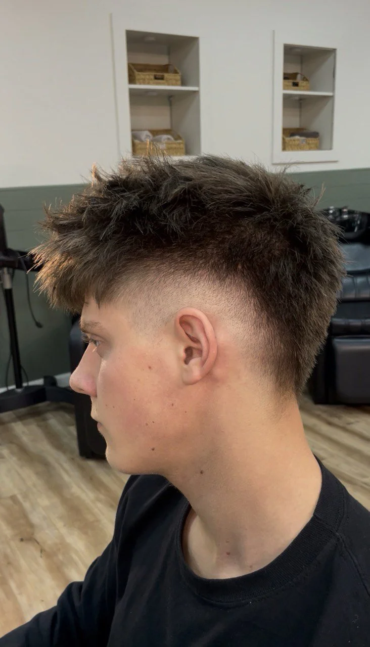 Side profile of a young man with a textured haircut featuring a fade on the sides and longer, tousled hair on top, sitting in a salon.