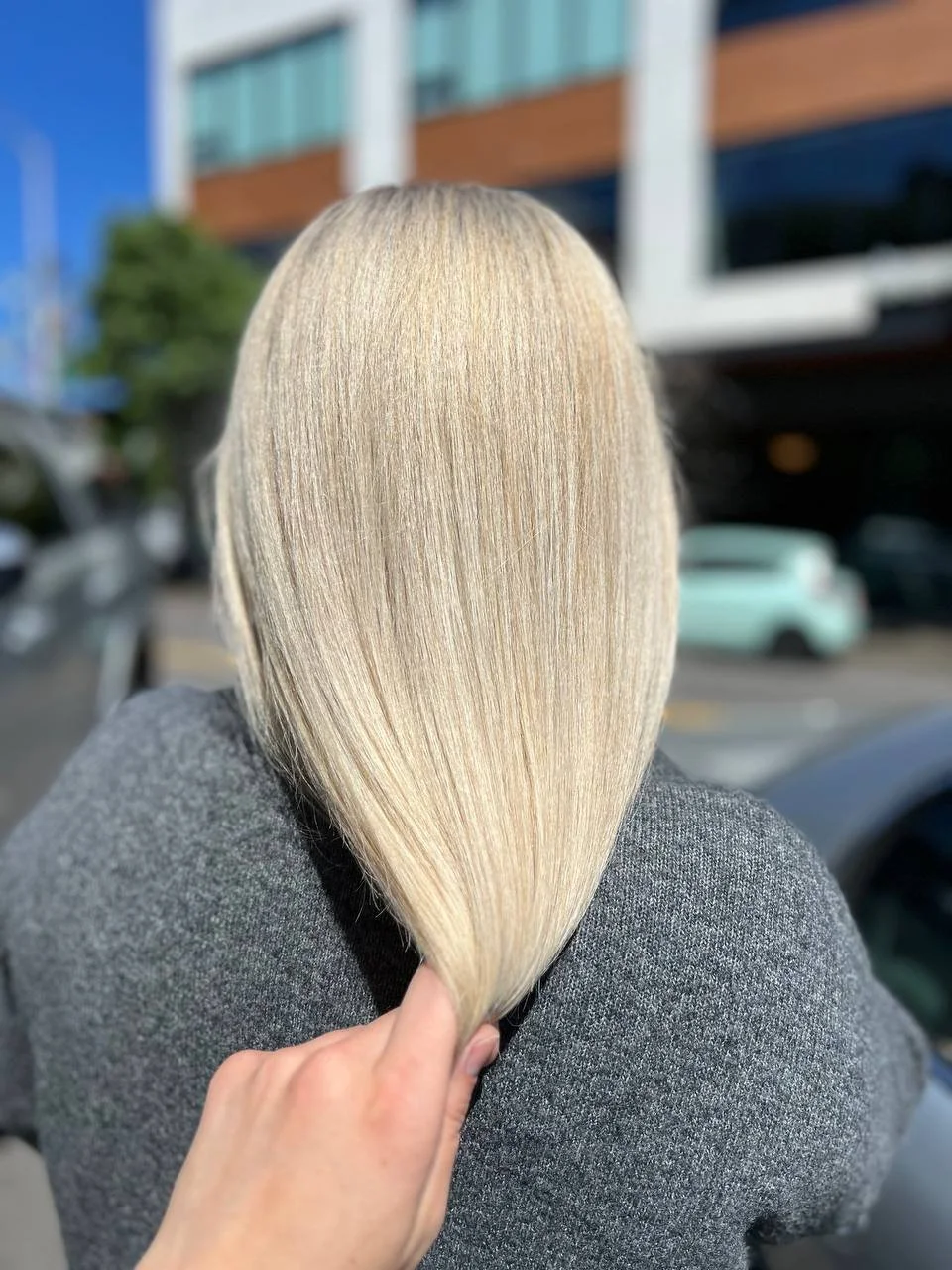 Person holding a strand of long, straight, blonde hair in front of their head outside with modern buildings, parked cars, and a tree in the background.
