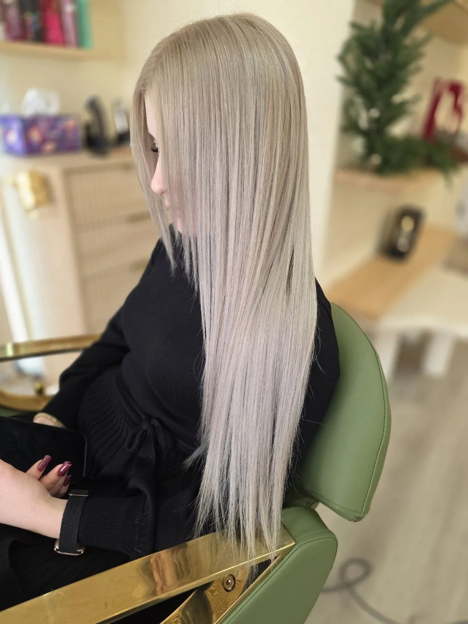 Woman with long, straight, platinum blonde hair sitting in a green salon chair, looking at her phone.