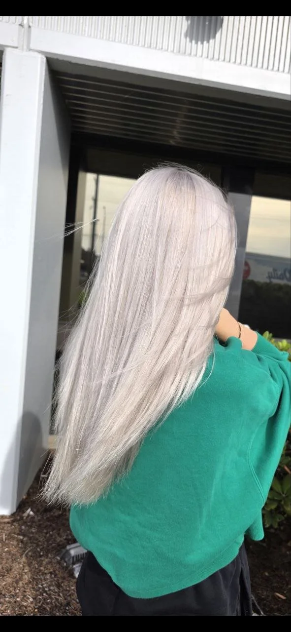 Person with long, straight, platinum blonde hair wearing a green shirt, standing outdoors near a building with a metallic exterior.