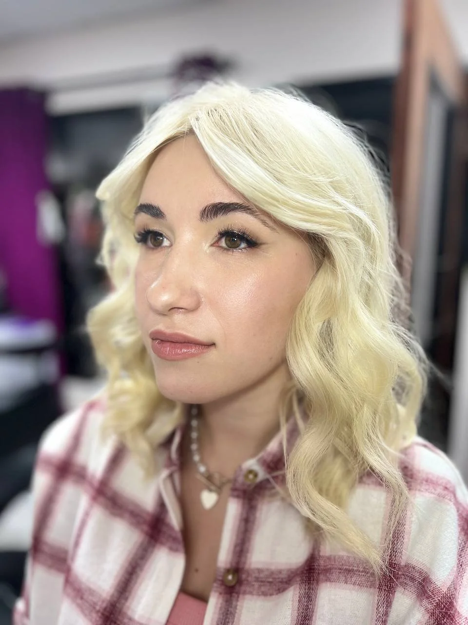 Close-up of a young woman with wavy platinum blonde hair, makeup, and jewelry in a salon or dressing room.