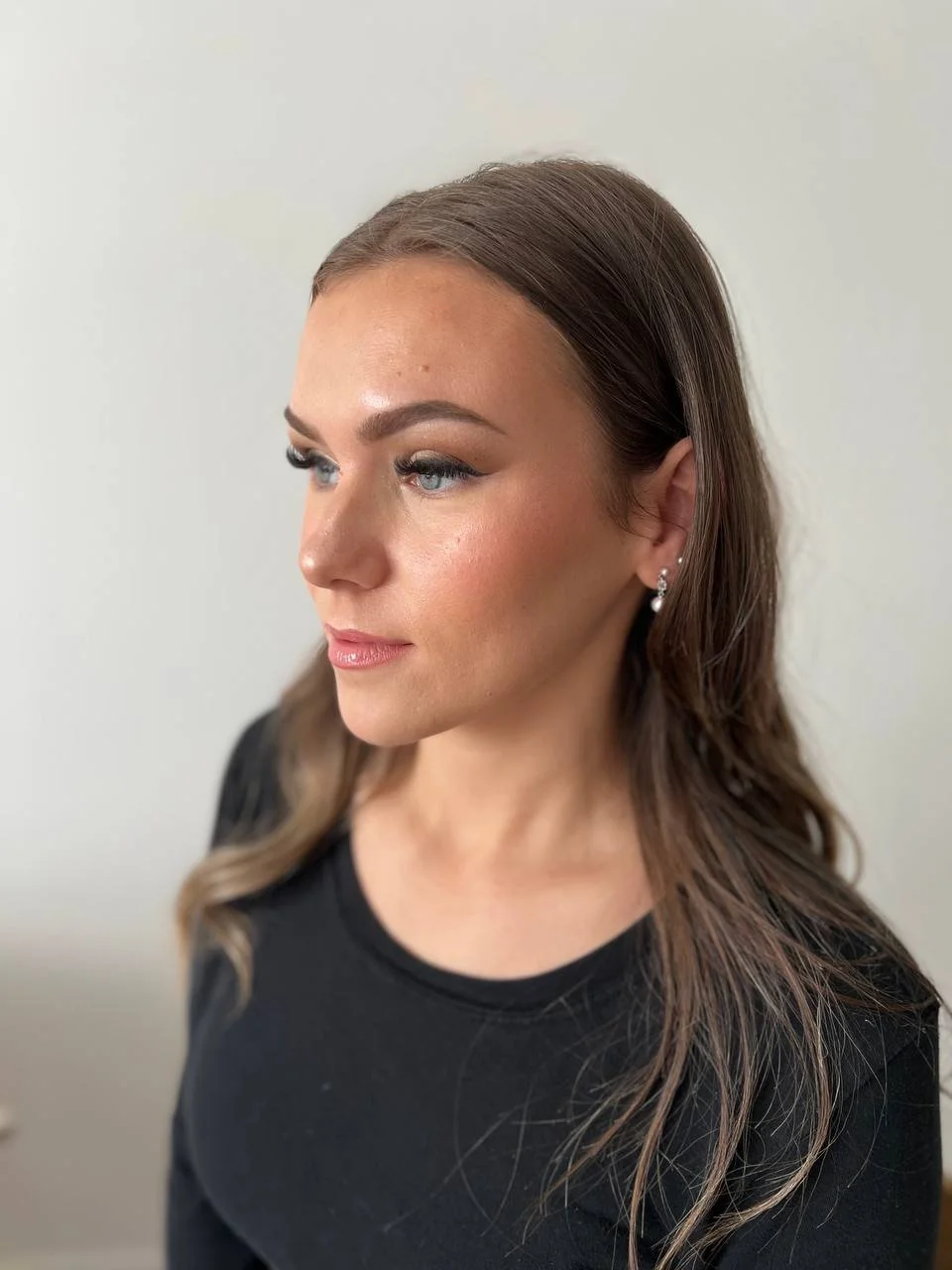 A woman with long, wavy brown hair, blue eyes, and makeup wearing a black top and earrings, looking to her left against a plain background.