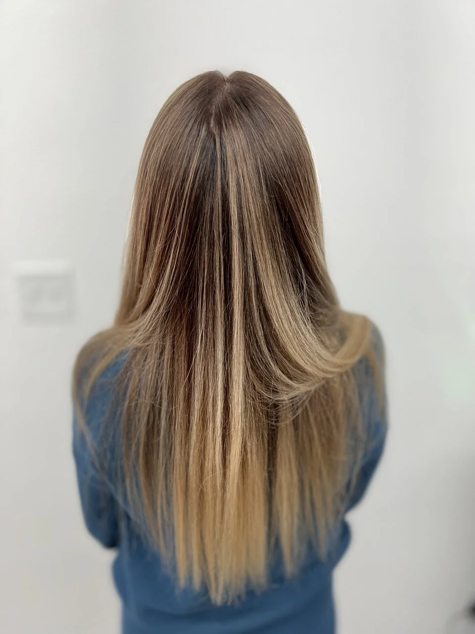Back view of a woman with long, straight, light brown hair with subtle highlights, wearing a blue top, standing against a plain light-colored wall.