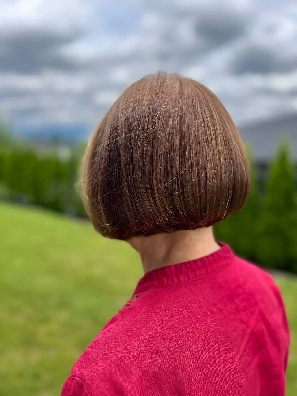 Back view of a woman with short, brown hair wearing a red top, outdoors with cloudy sky and green trees in the background.