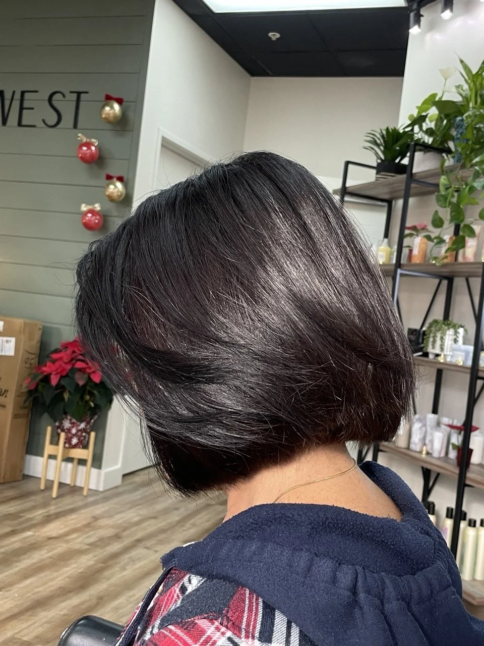 A woman with a sleek, shiny, dark bob haircut sitting in a salon with holiday decorations, green plants, and shelves with products in the background.