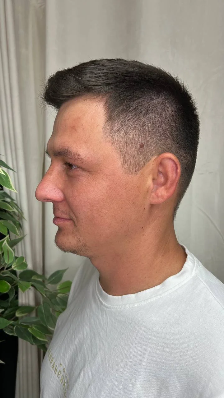 Profile side view of a man with short, dark hair, wearing a white T-shirt, standing next to a green leafy plant with beige curtains in the background.