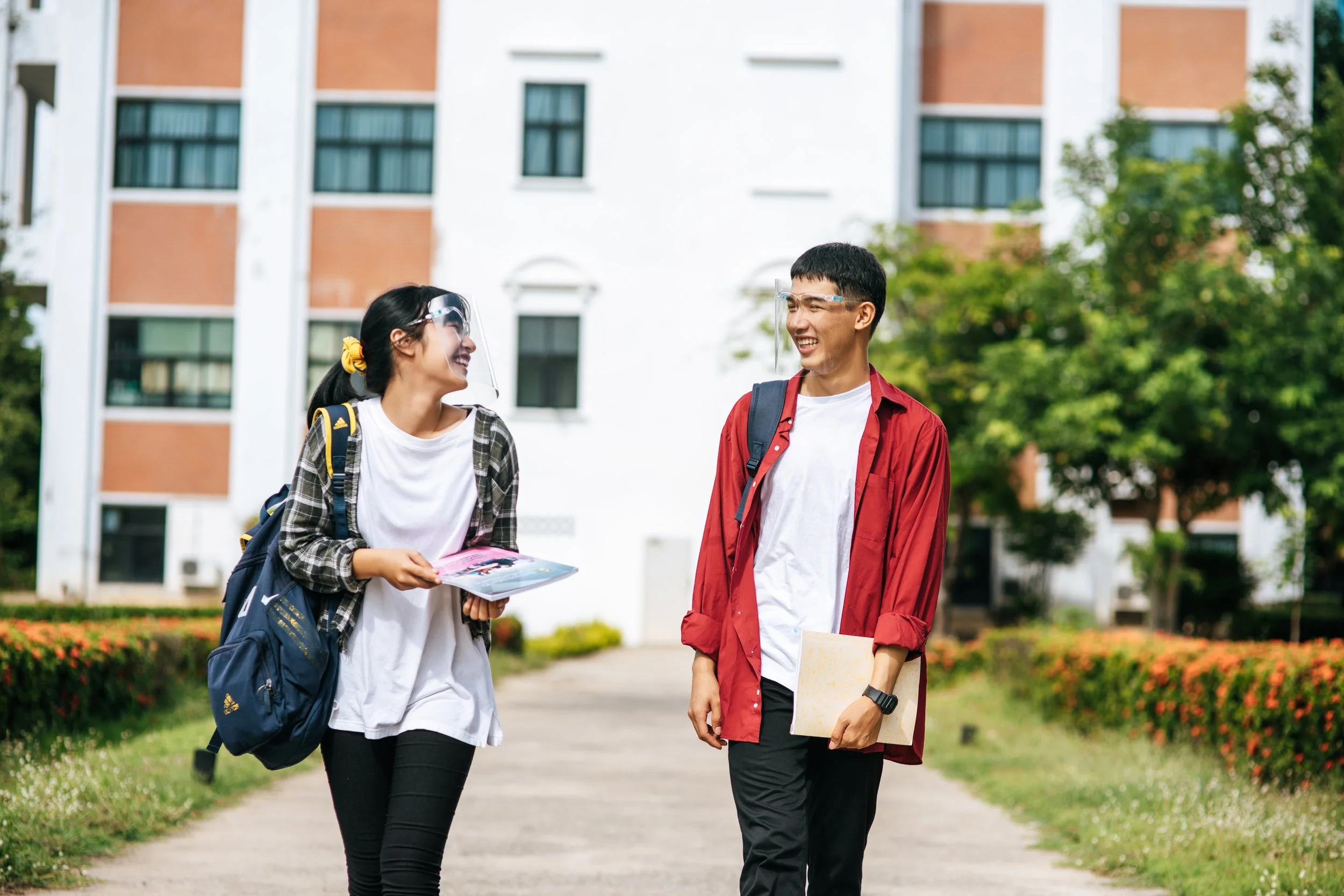 Two students walking outside on a university campus, wearing face shields and backpacks, smiling and talking to each other.
