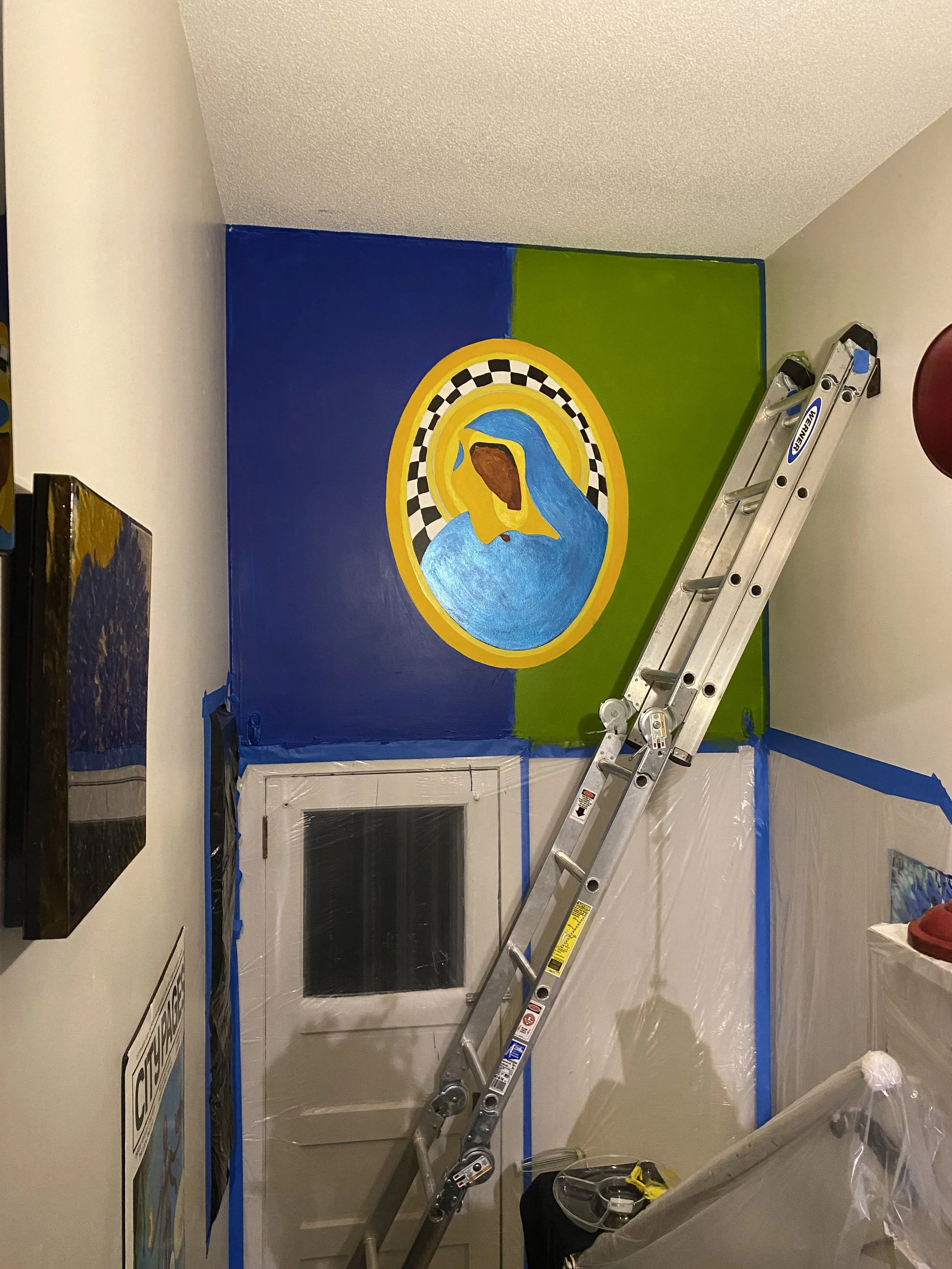 A mural in progress above a doorway showing an early stage of a Black Madonna figure draped in blue, set within a gold halo with a checkered border, against a split blue and green background.