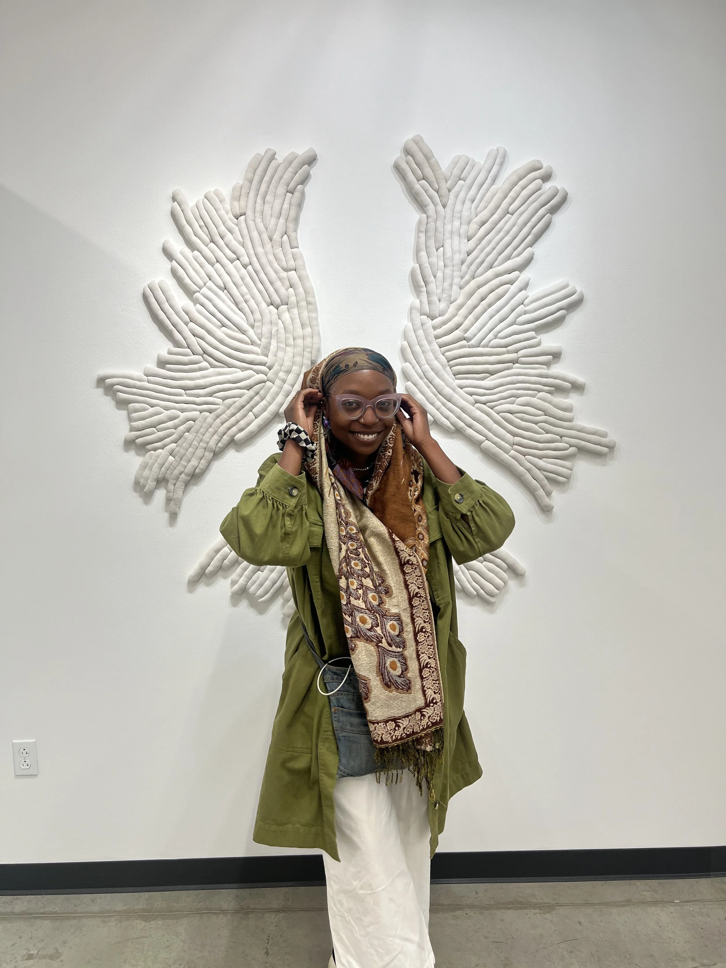 Haitian-American artist Raph Draws smiling in front of a large white sculptural wing installation, wearing a green jacket, patterned scarf, and lavender cat-eye glasses
