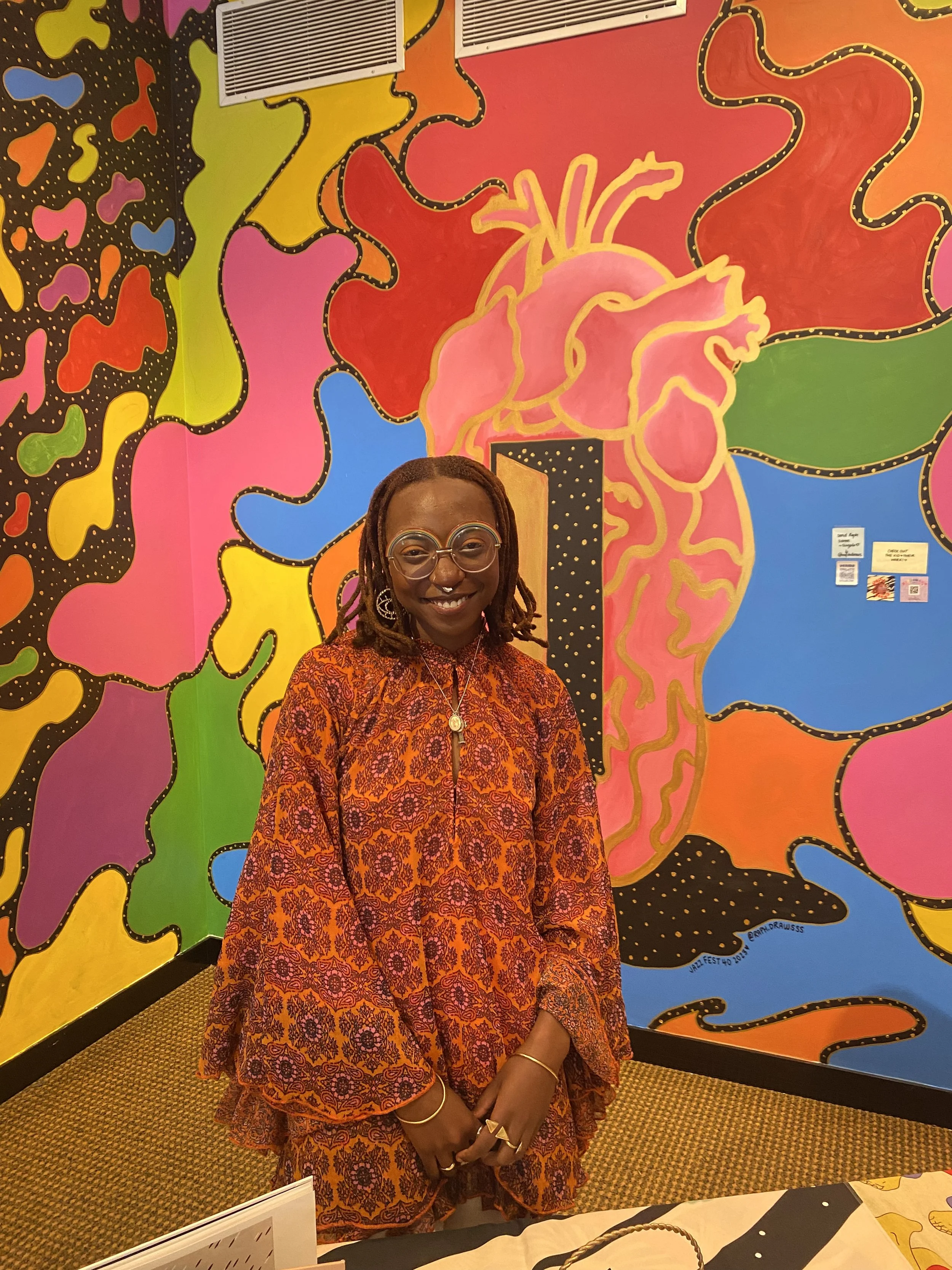 Haitian-American artist Raph Draws smiling in front of her Journey Through Vibe N' Flow collaborative mural at Burlington Discover Jazz Fest 40, wearing an orange patterned dress and round silver glasses