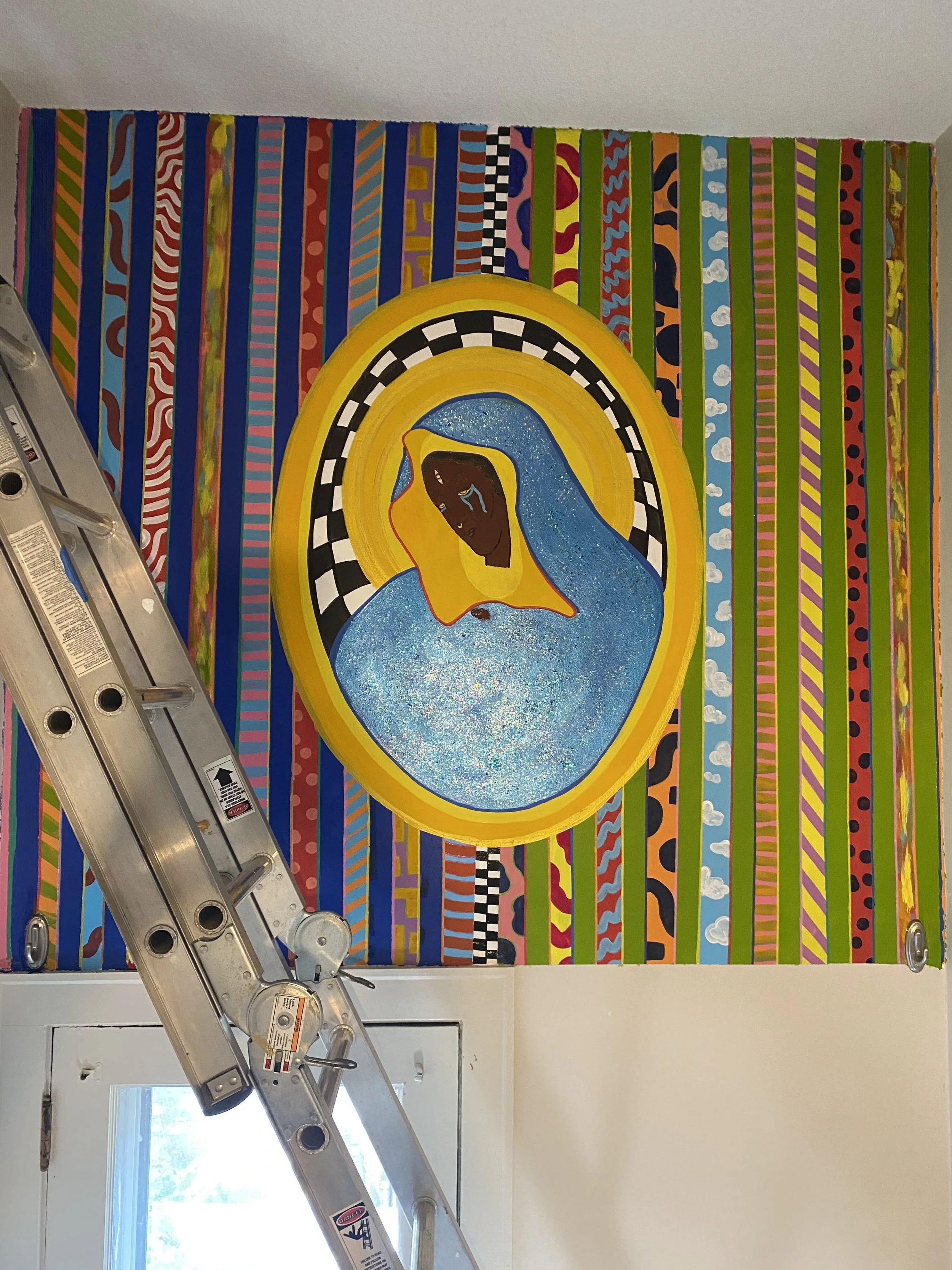 A wide view of a mural in progress showing the full wall of vertical patterned stripes in blue, green, orange, and red, framing a Black Madonna figure in a gold halo centered above a doorway.