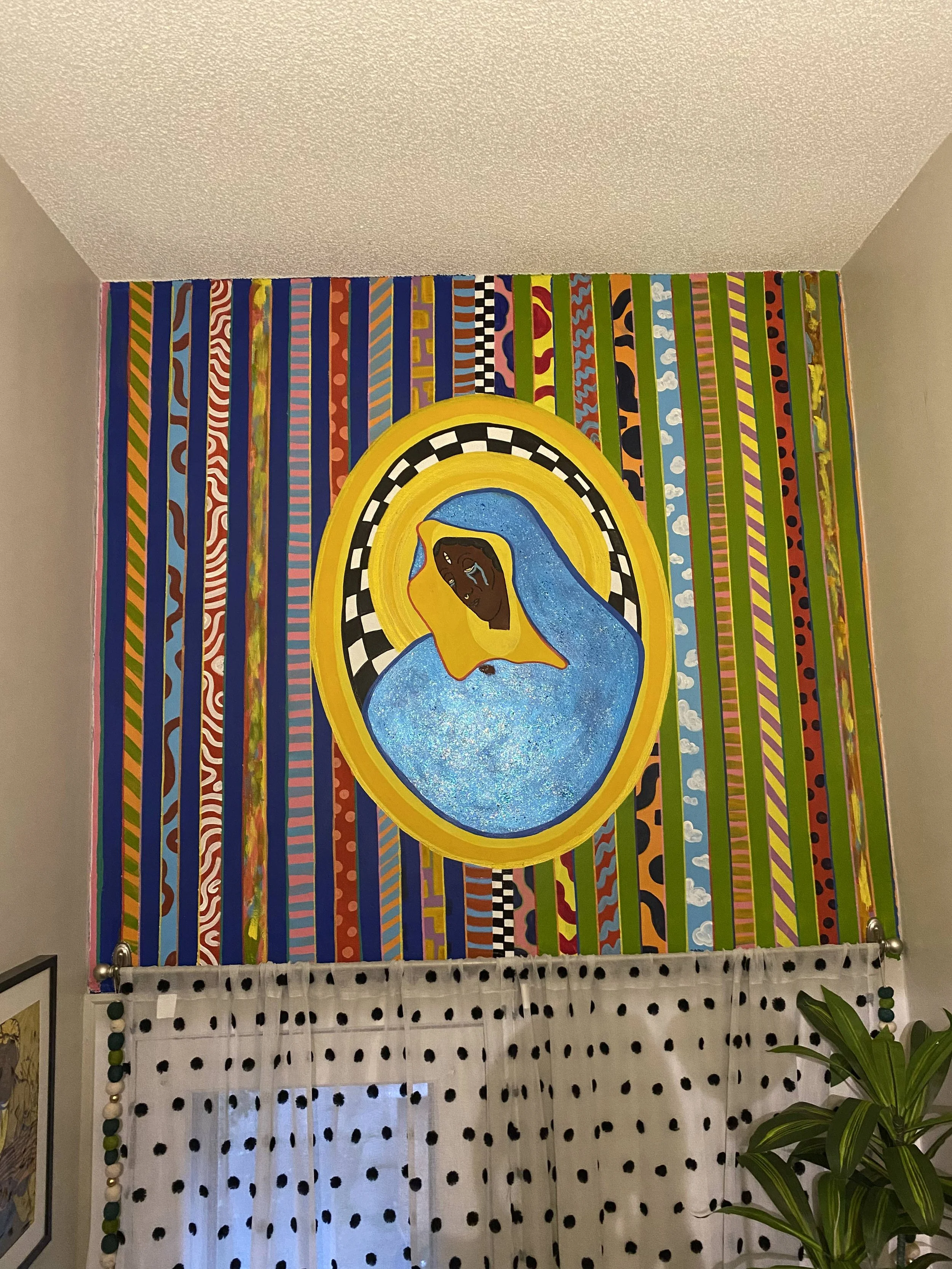 The completed mural showing a weeping Black Madonna with a dark face and shimmering blue robe, set within a gold oval halo with a black and white checkered border, centered on a wall of richly patterned vertical stripes.