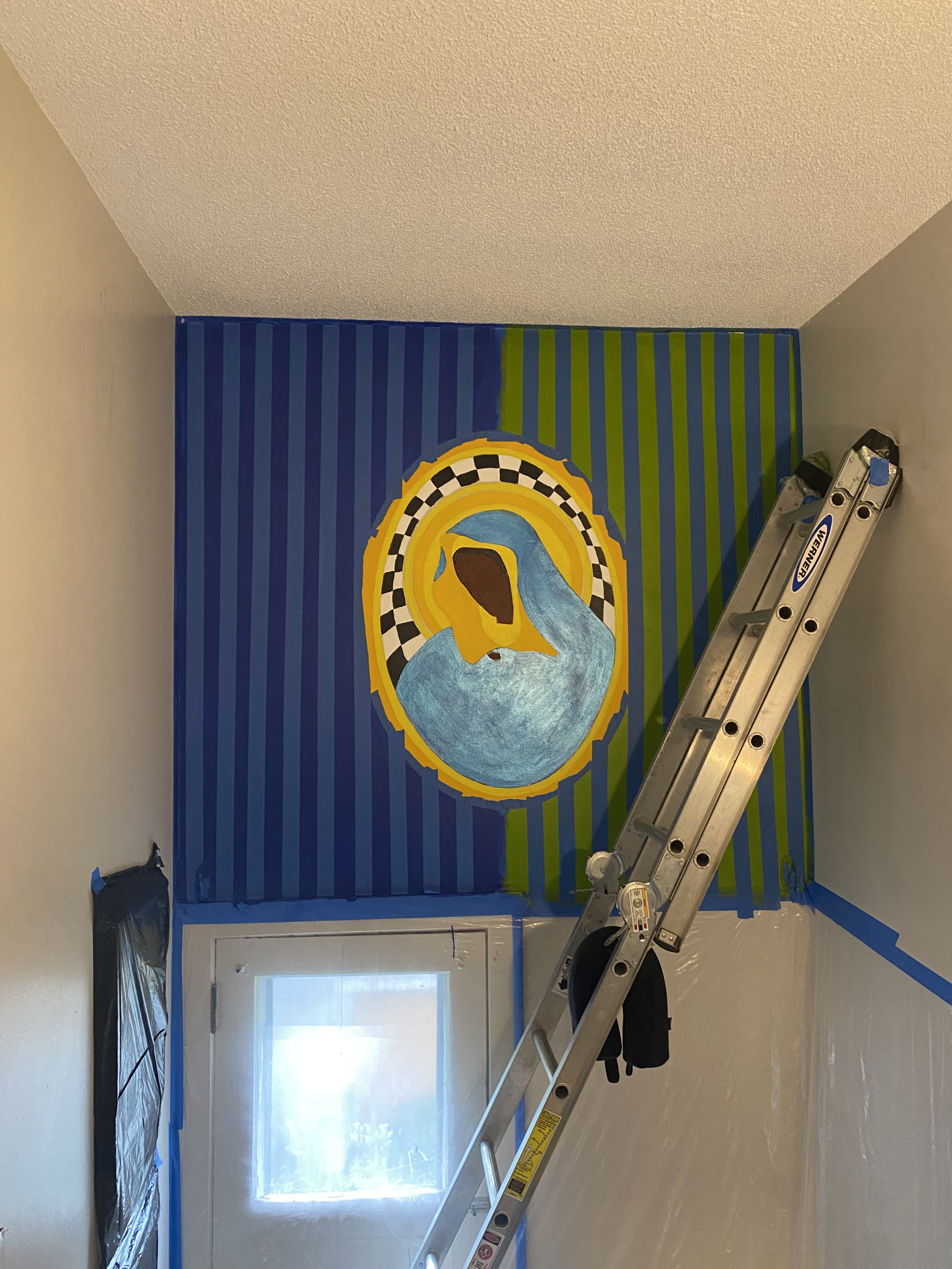 A mural in progress showing a Black Madonna in a gold oval halo above a doorway, now surrounded by vertical blue and green stripes painted across the wall.